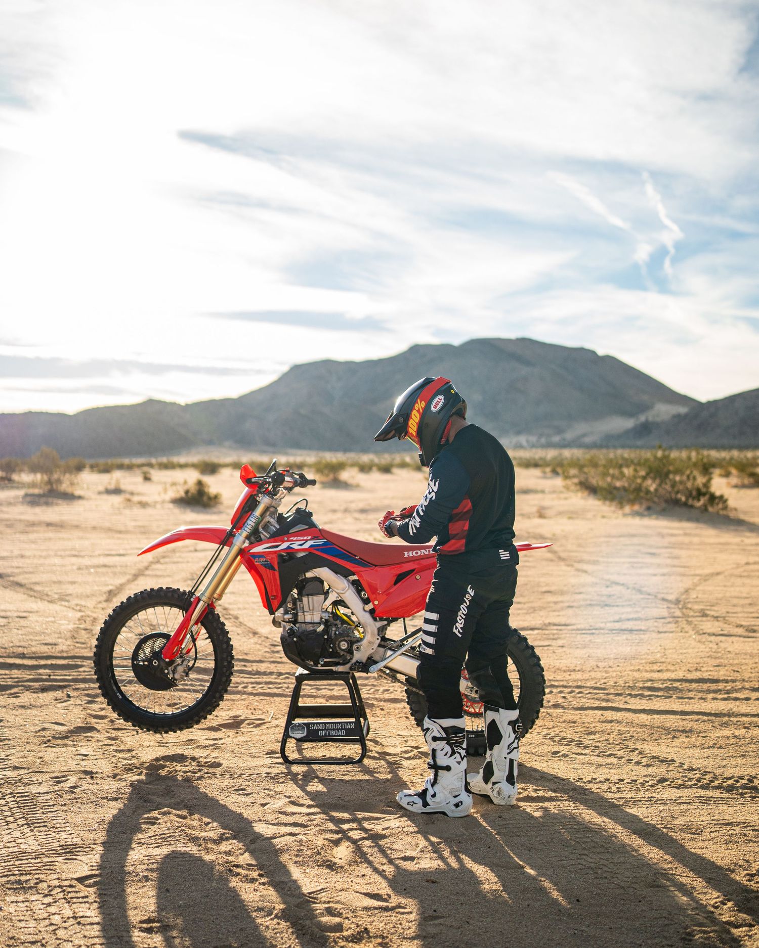 A dirt bike rider performs maintenance on a red motorcycle in a desert landscape with mountains in the background.