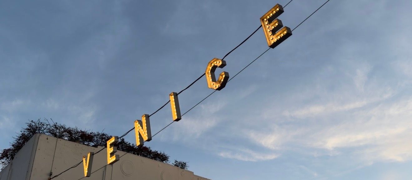 Vintage neon letters spelling 'VENICE' mounted on a wire against a cloudy blue sky.