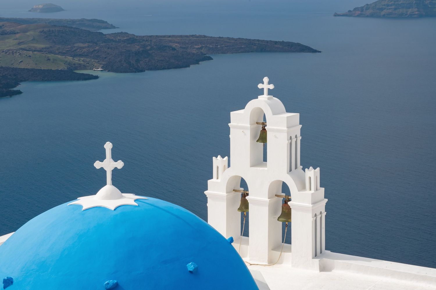 White Greek Orthodox churches with blue domes overlook the stunning Aegean Sea and volcanic islands of Santorini.