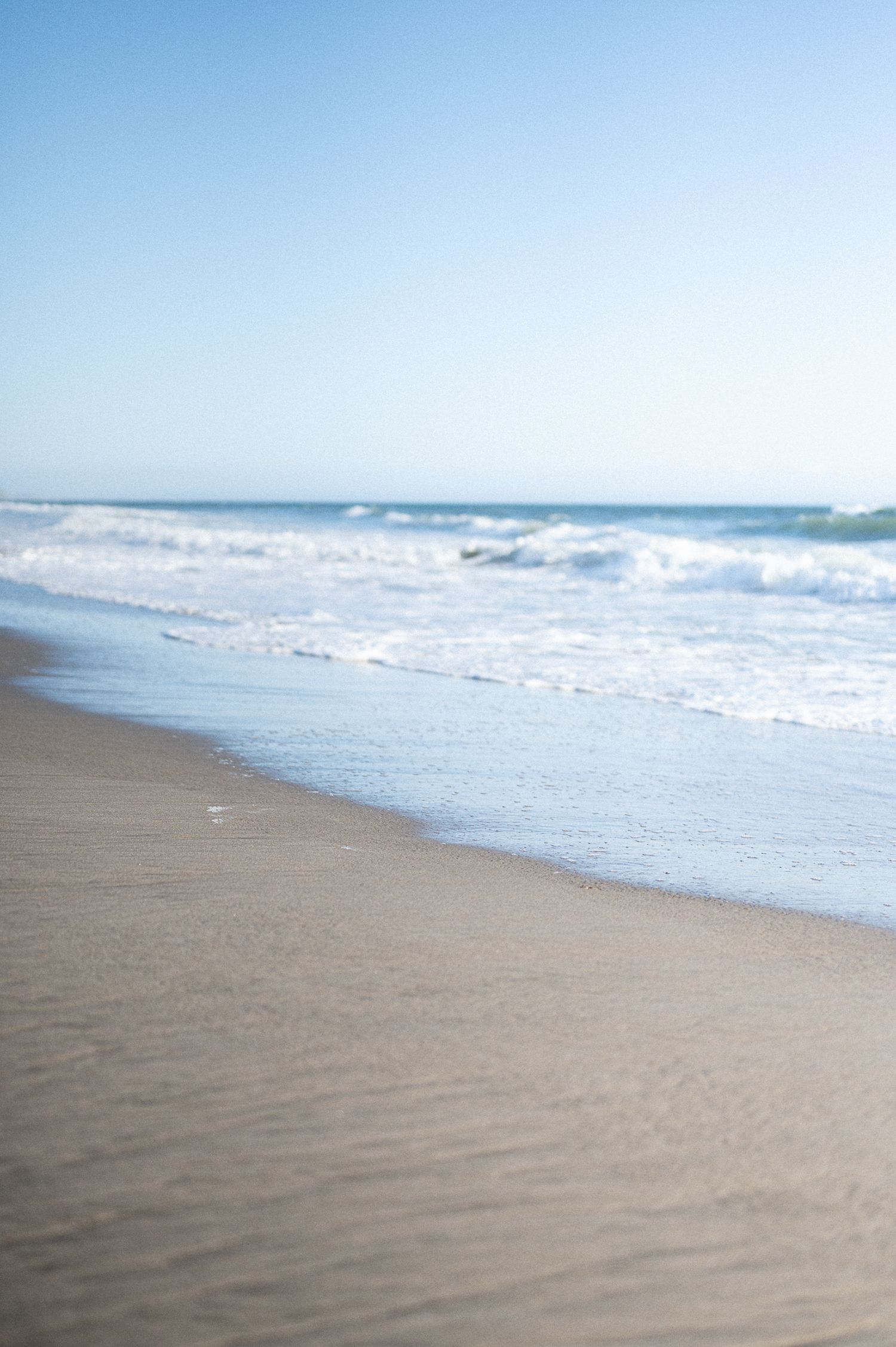 Serene beach scene with gentle ocean waves washing onto smooth sandy shore under a clear blue sky.
