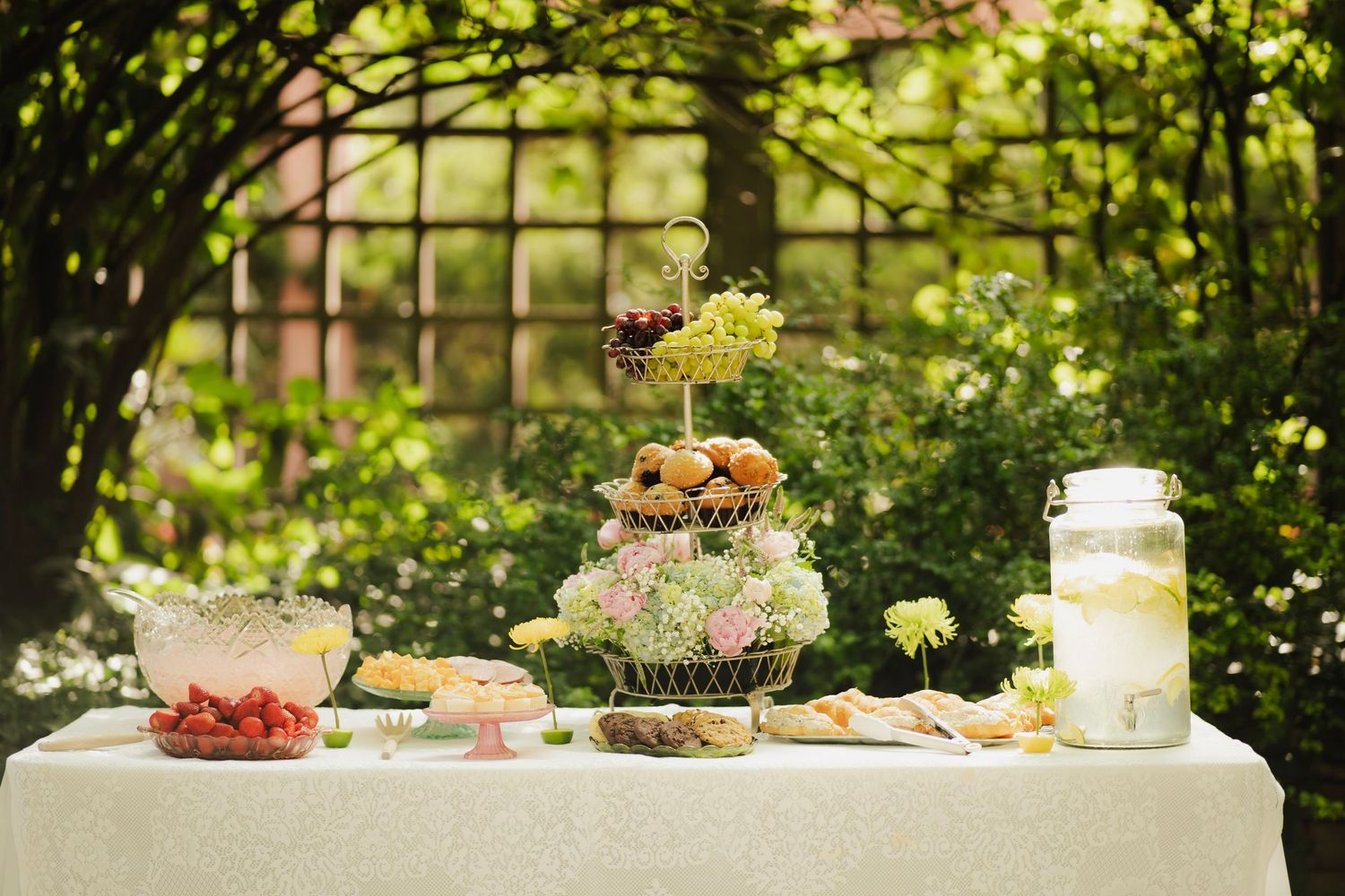 Outdoor garden party dessert table with tiered fruit display, fresh flowers, and lemonade dispenser.