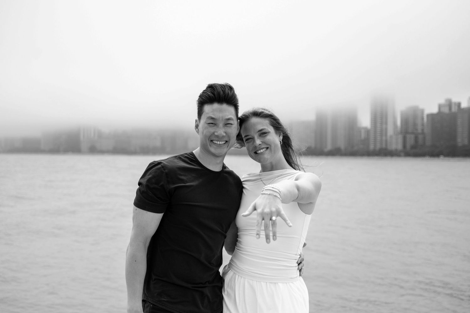 Happy couple poses by waterfront with city skyline, woman showing engagement ring, misty urban backdrop.