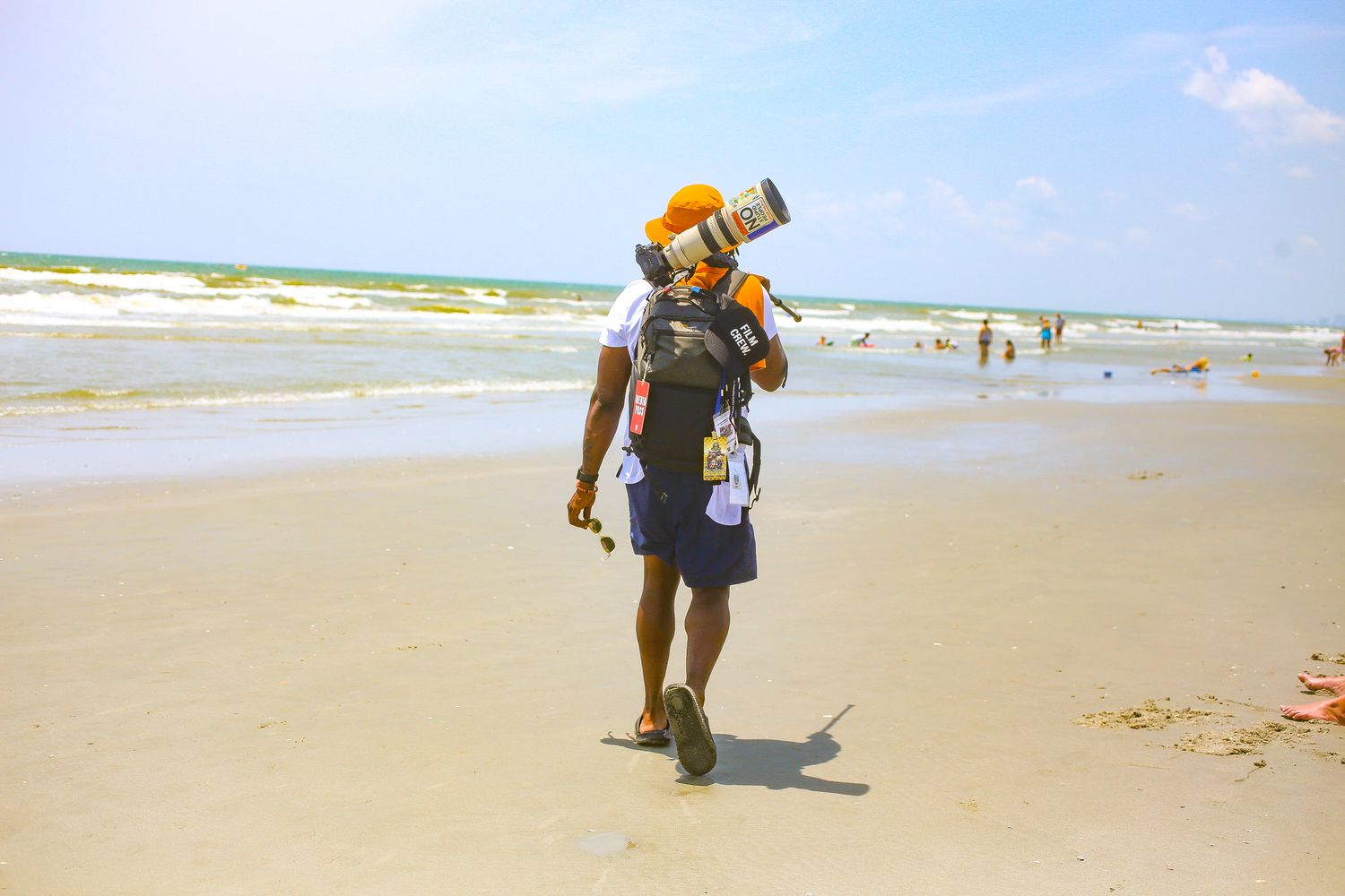 Photographer with large camera lens walks on sunny beach with ocean waves and beachgoers in background.