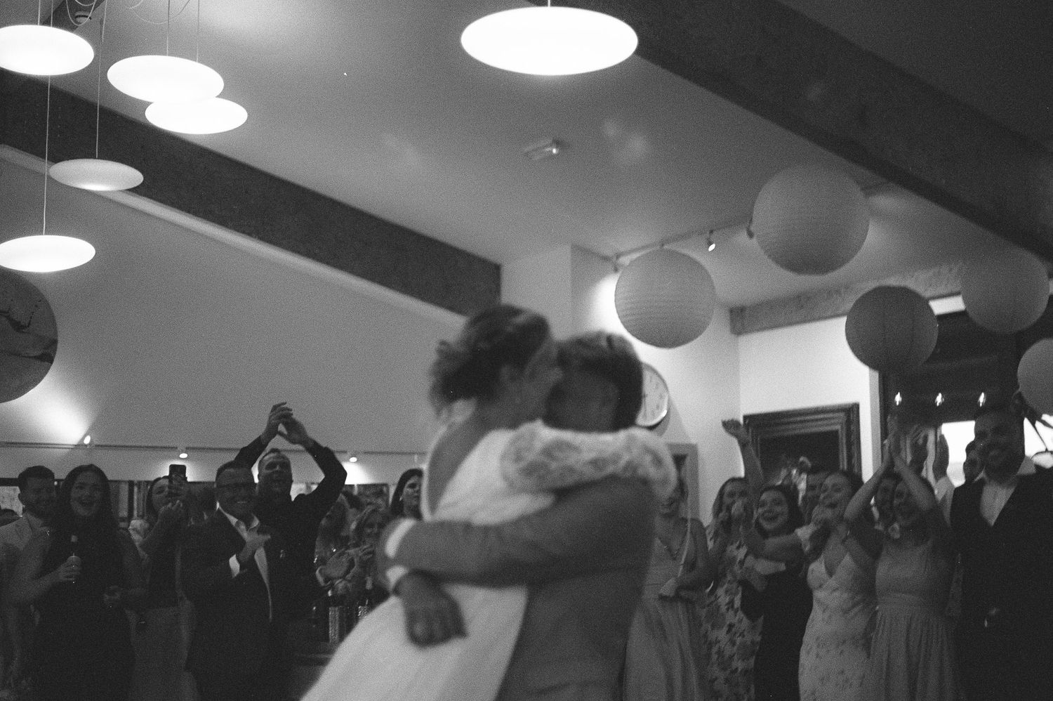 Black and white photo of couple sharing first dance at wedding reception with guests celebrating in background.