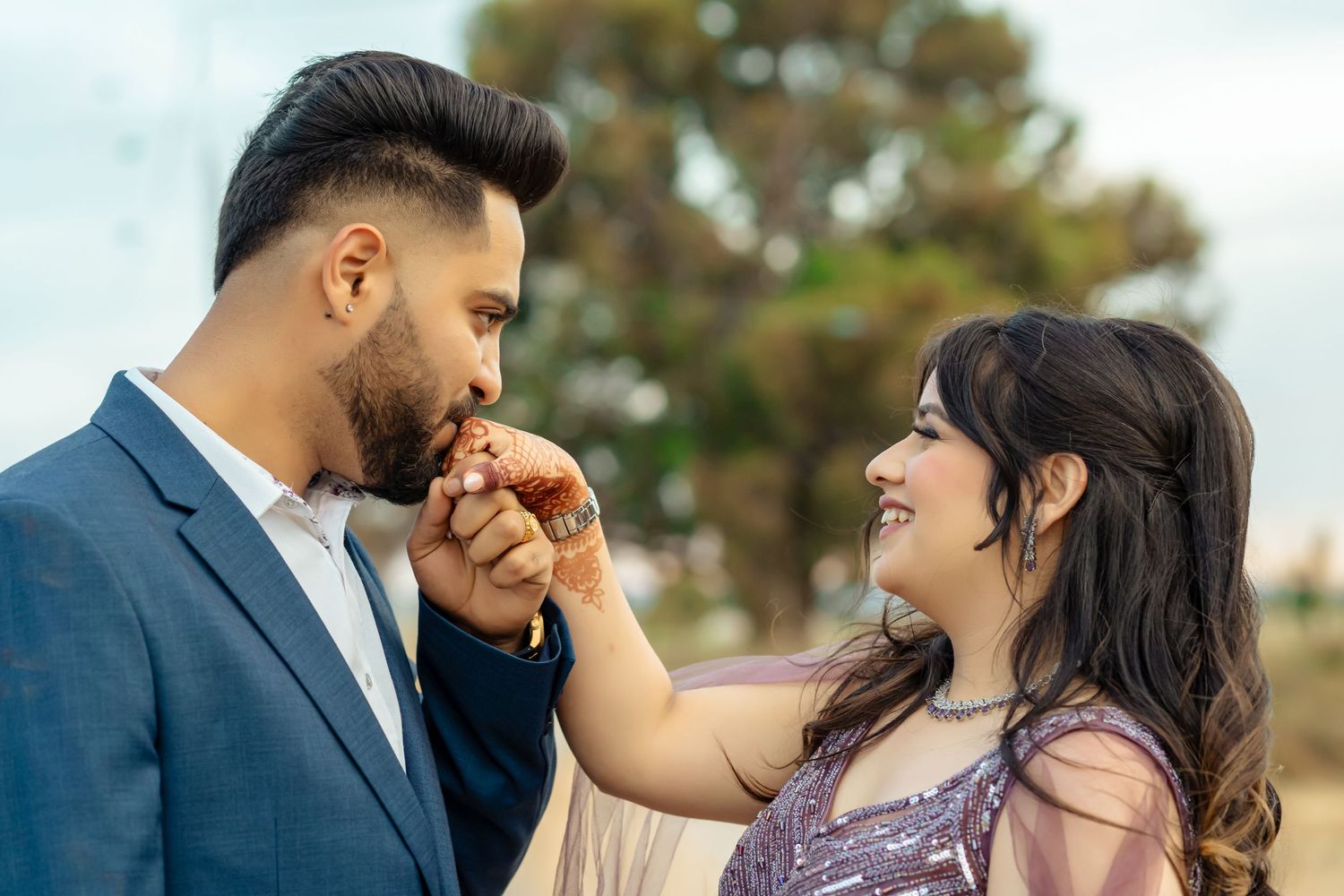 Couple in elegant Indian attire, woman in golden lehenga with jewelry, man in brown blazer, posing at a formal event.