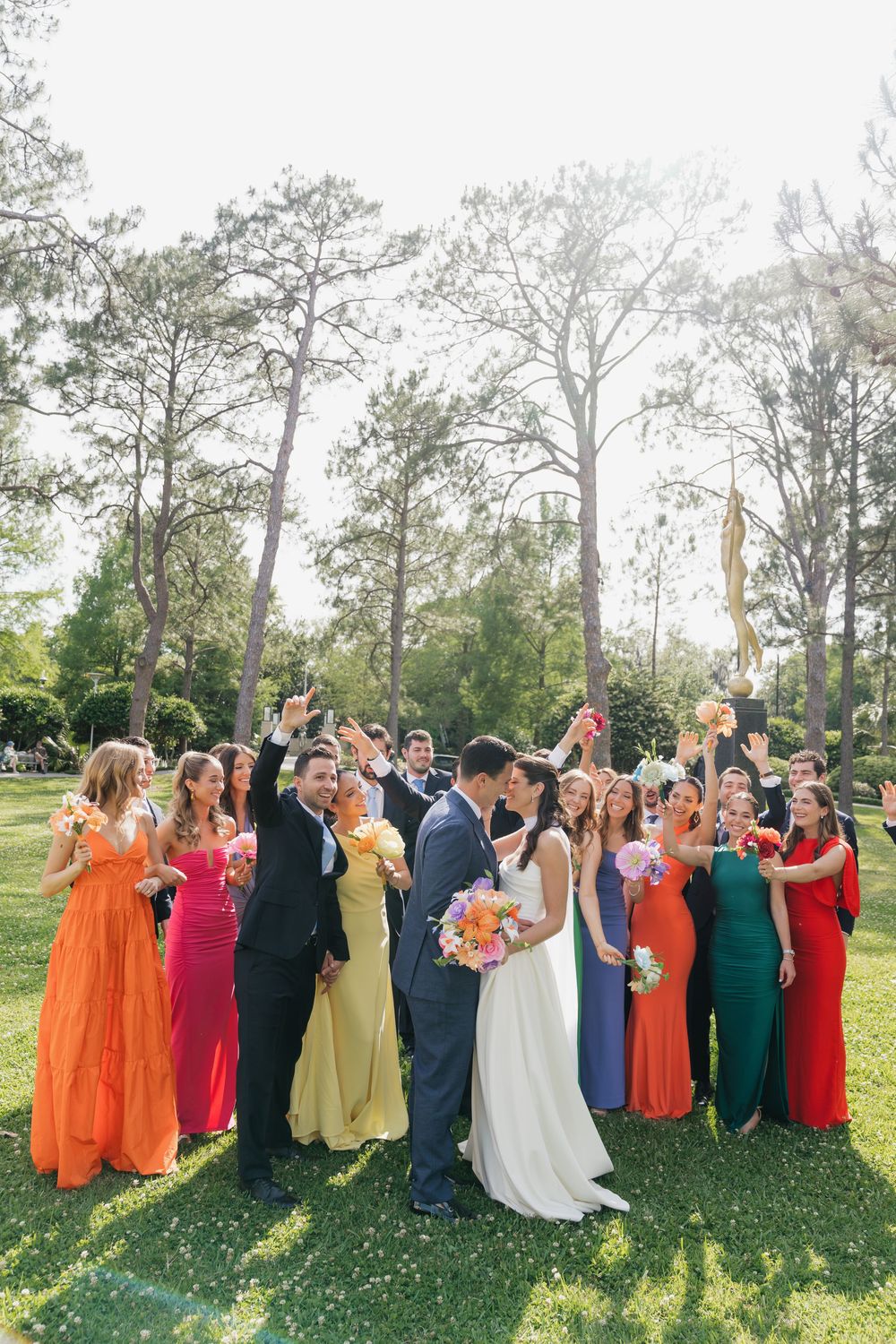Wedding party celebrates outdoors with bride and groom kissing, bridesmaids in colorful dresses raising glasses.