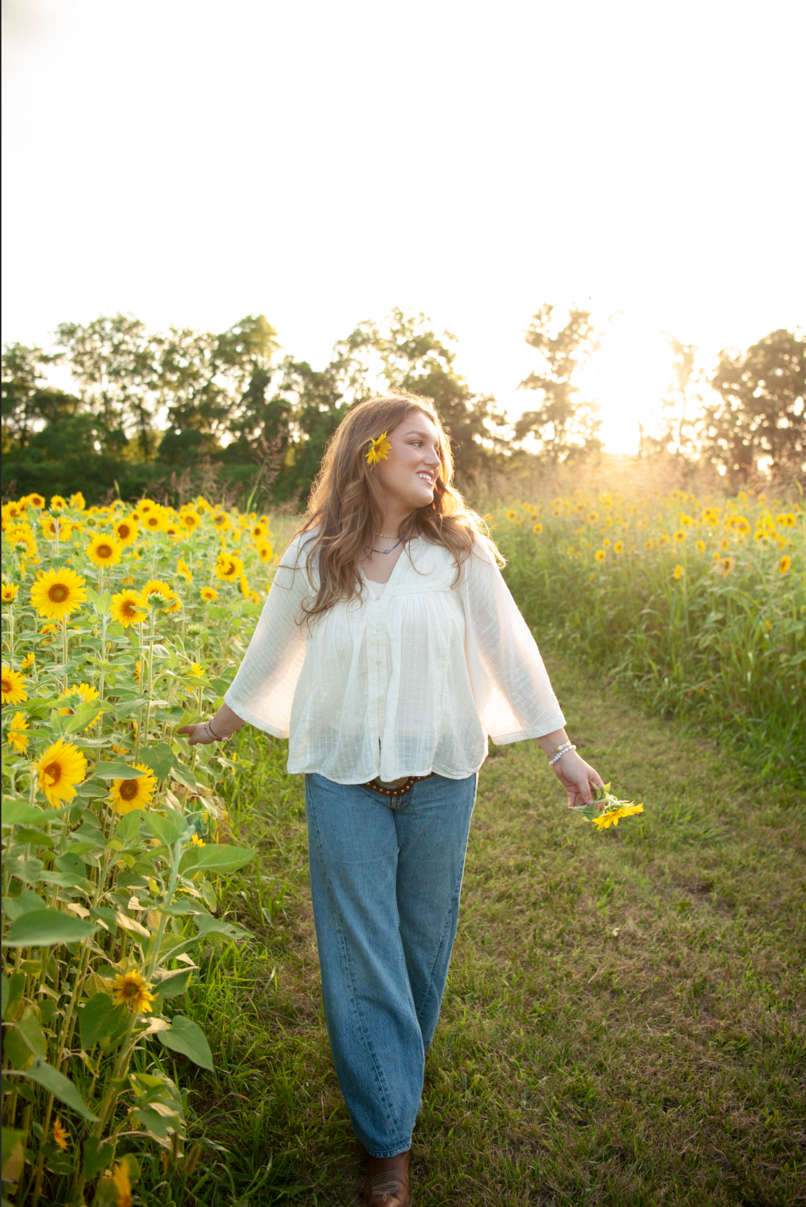 Young woman in white blouse and jeans walks through a sunflower field at golden hour, holding yellow flowers.