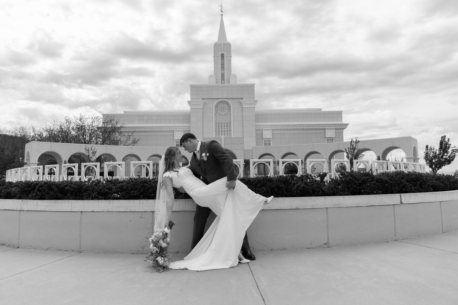 Black and white photo of a groom dipping his bride in front of a grand white LDS temple on their wedding day.