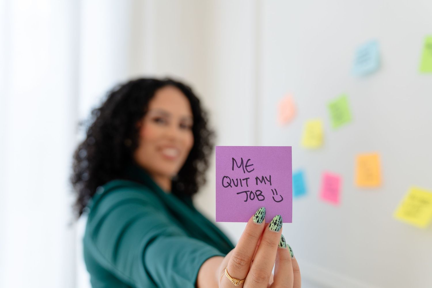 A person in a teal sweater holds colorful sticky notes while working at a desk with a laptop and office supplies.