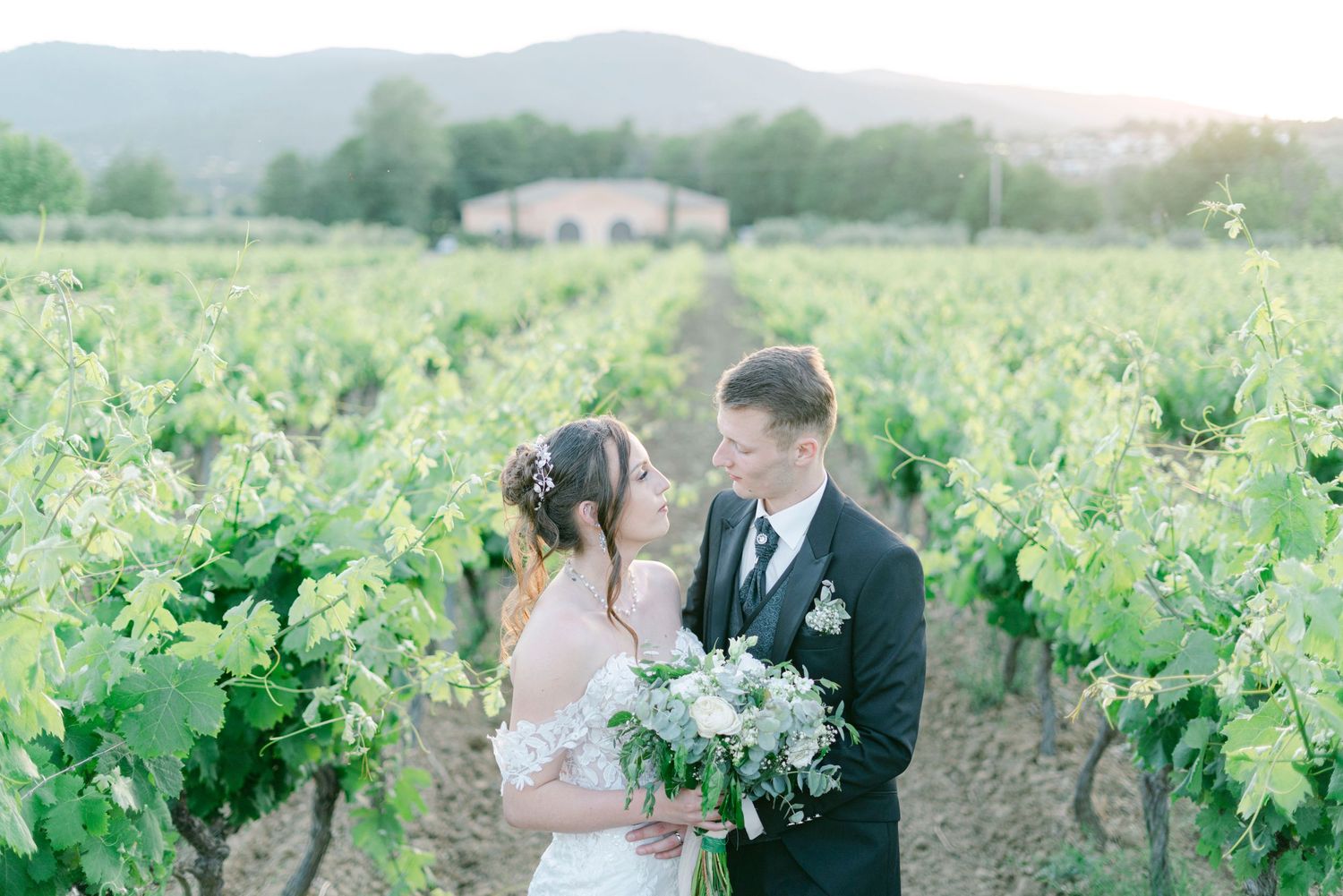 A romantic couple embraces in a lush vineyard setting during a wedding photoshoot at sunset.