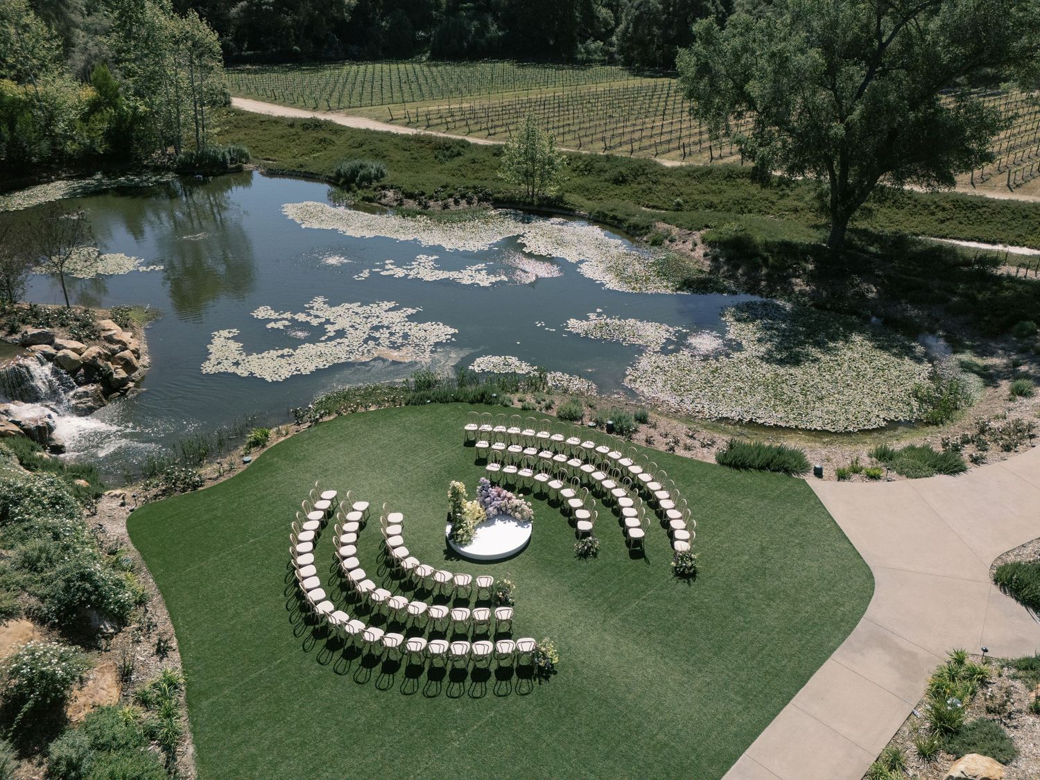 Aerial view of an outdoor wedding ceremony setup with white chairs arranged in semi-circles near a decorative pond.