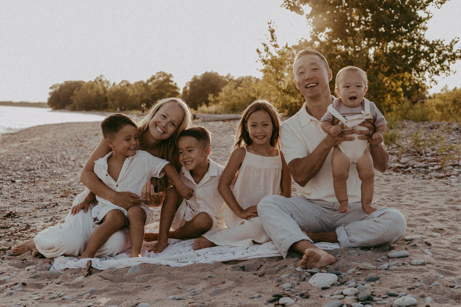 Happy family of six sitting together on sandy beach at golden hour, parents with four young children.