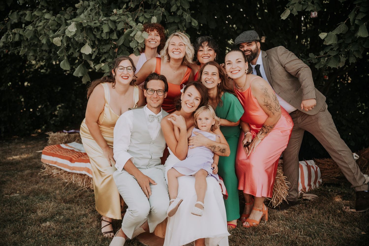 A joyful group of wedding guests posing together outdoors on hay bales, wearing colorful formal attire in a garden setting.