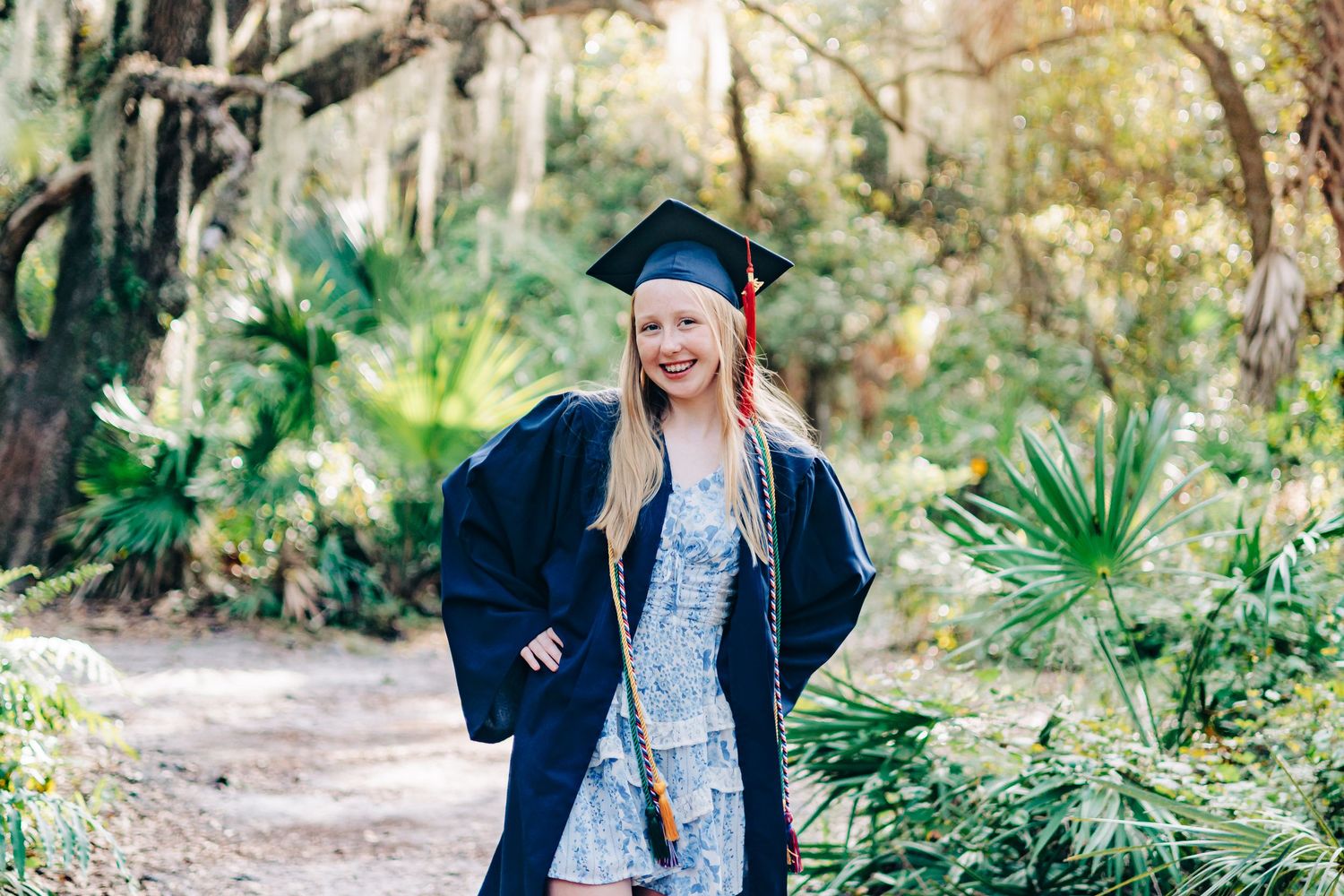 Smiling graduate in cap and gown poses on a scenic forest path surrounded by lush green trees and palms at Delray Oaks Natural Area.