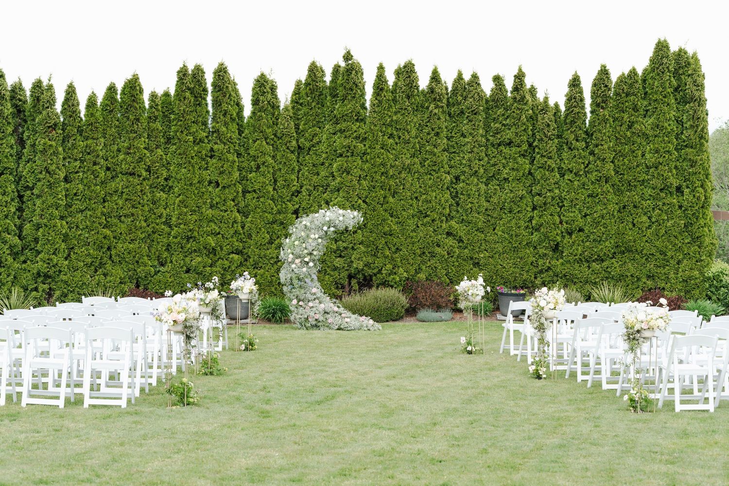 Outdoor wedding ceremony setup with white chairs, floral crescent arch, and tall green arborvitae hedge backdrop.