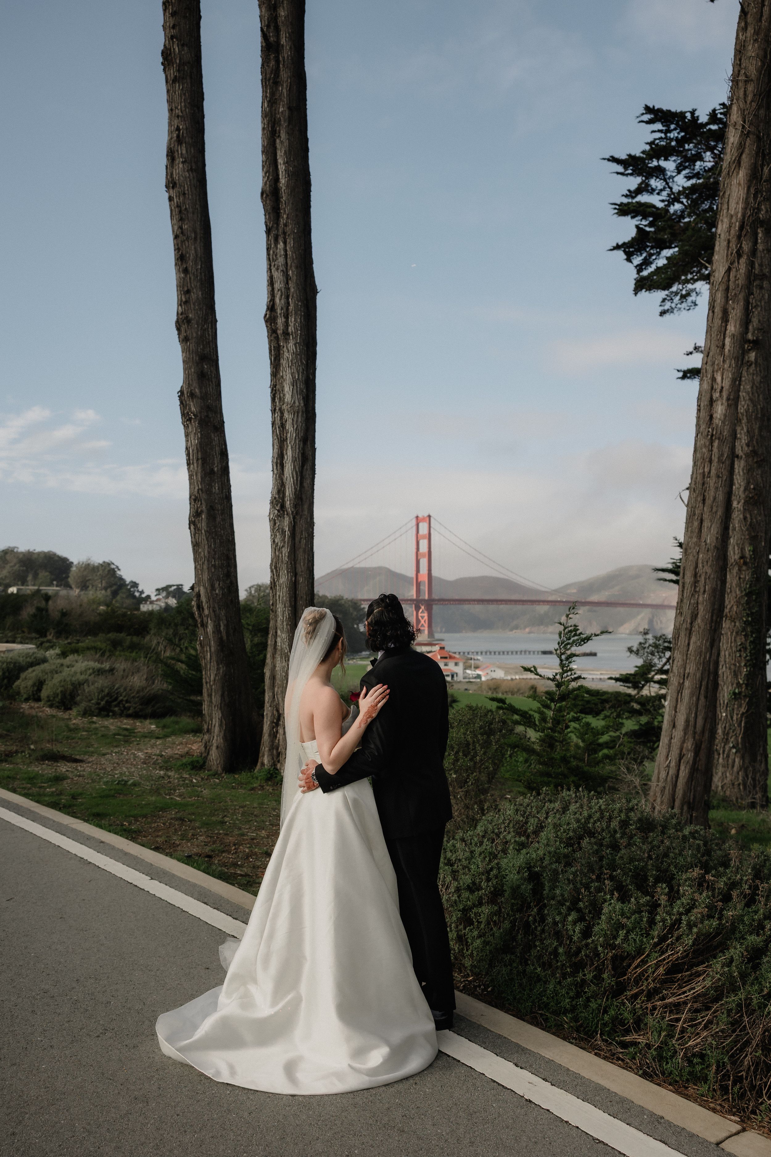 Bride and groom embracing near Golden Gate Bridge in San Francisco, surrounded by tall trees in a romantic outdoor wedding portrait.