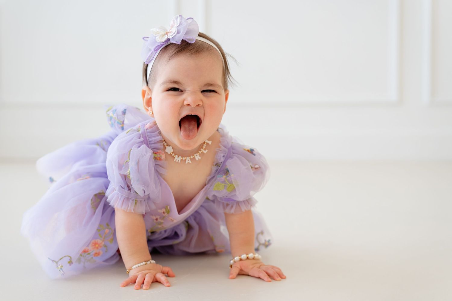 Baby girl in lavender tutu dress and pearl jewelry crawls on white floor, mouth open in joyful expression.
