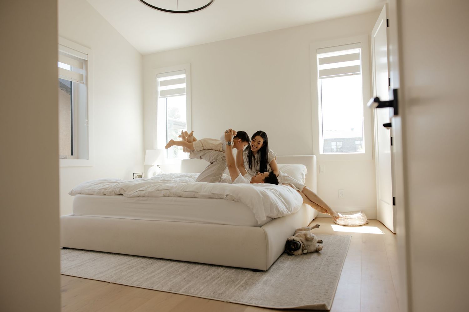 Two women playing on a white bed in a bright, modern bedroom with large windows and a dog resting on the rug.