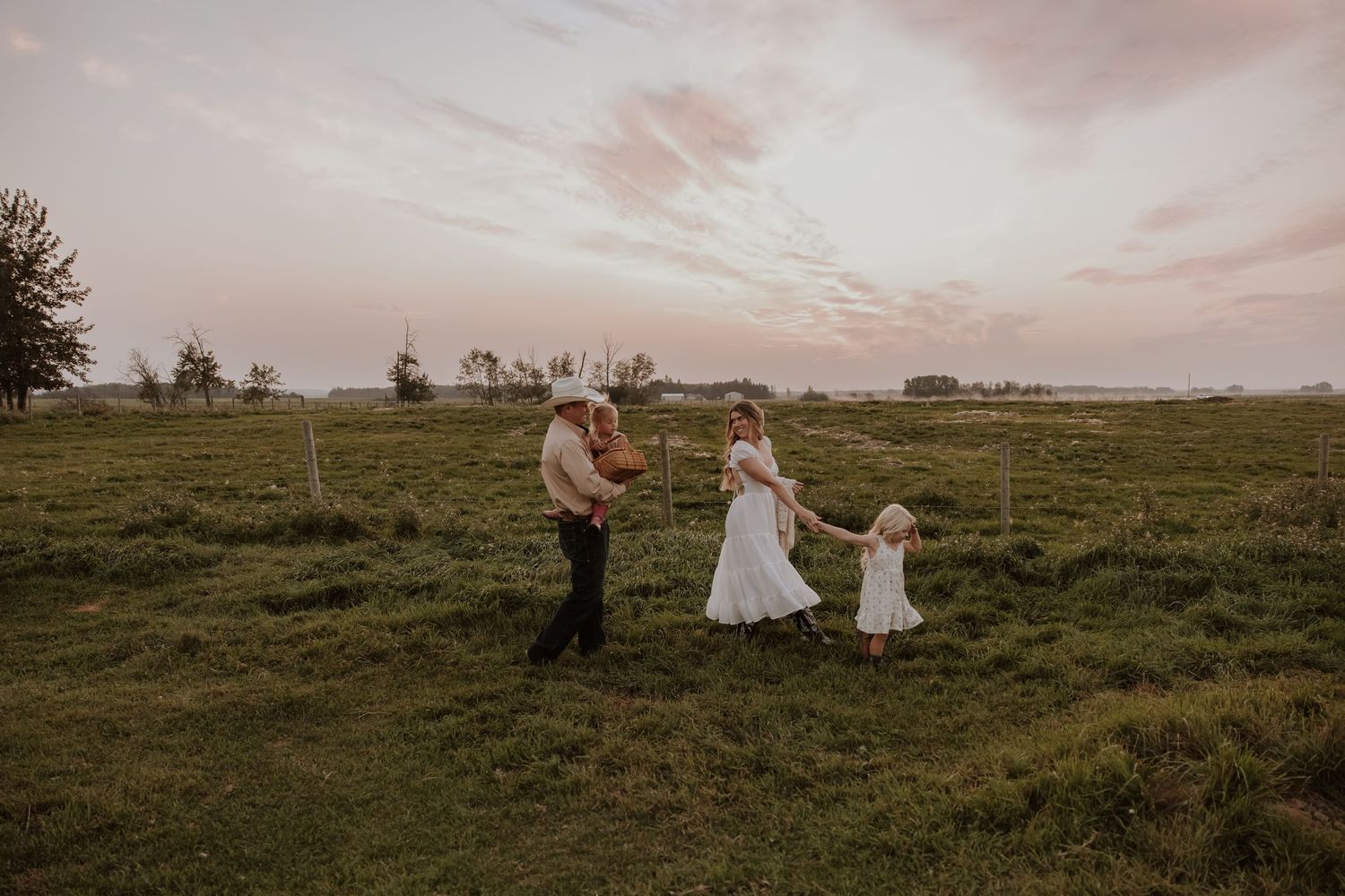 Family of three walking in green field at golden hour sunset, parents holding toddler girl's hands in white dress.