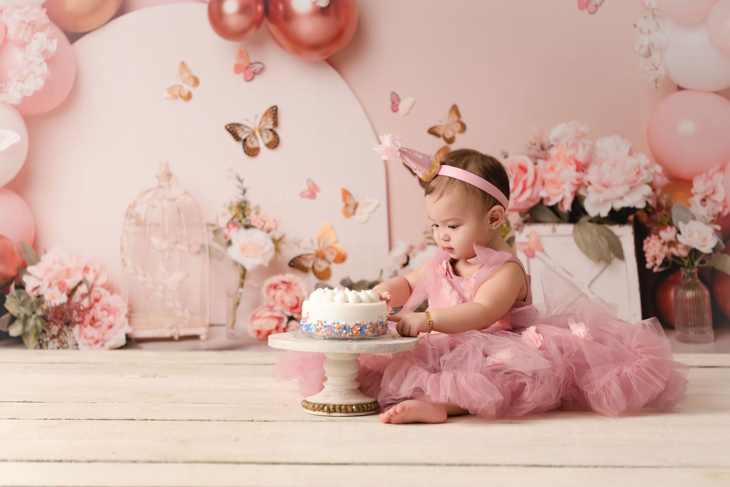 Baby girl in pink tutu smashes first birthday cake surrounded by pink balloons, flowers, and butterfly decorations.