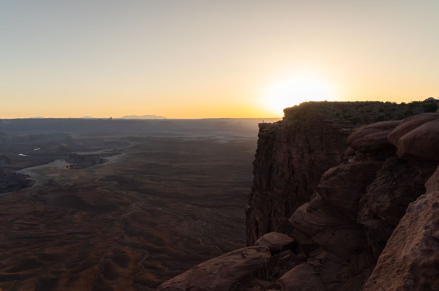 A figure stands on a cliff edge, surrounded by a panoramic sunset scene in Canyonlands National Park, Utah.