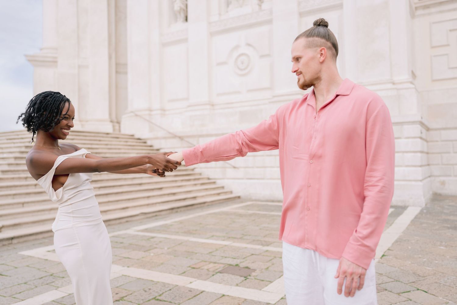 A joyful interracial couple holds hands outdoors, dressed in elegant white and pink outfits near a grand white building.