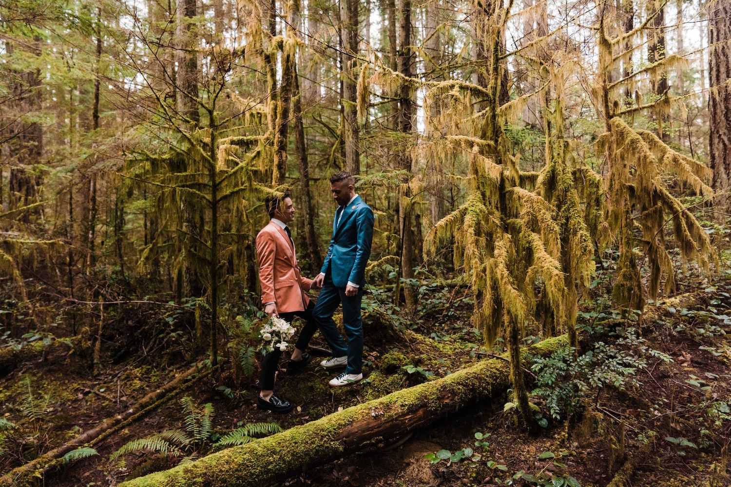Two people in colorful suits walk on a mossy log in a lush, green forest during an outdoor elopement.