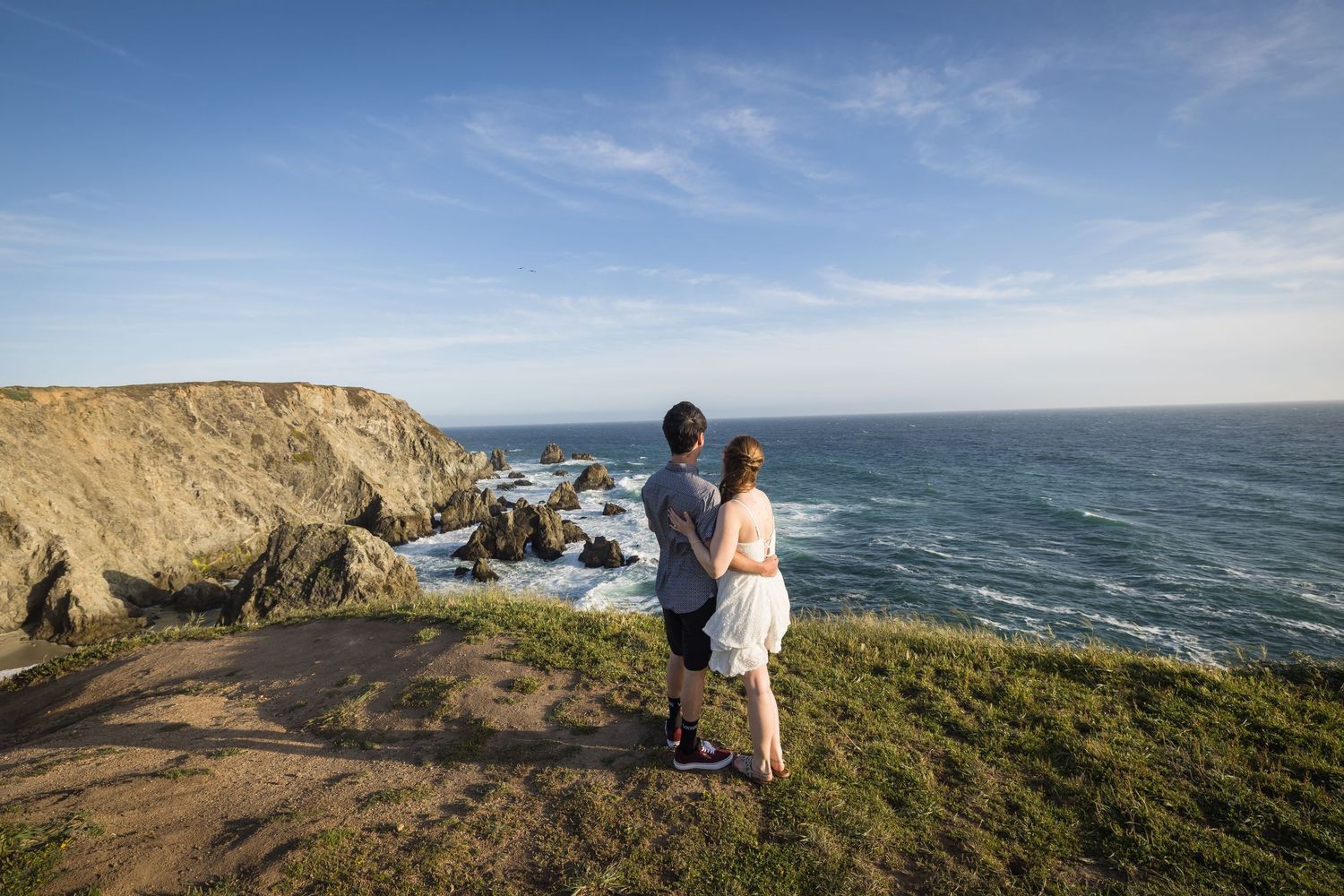Couple standing on a coastal cliff overlooking rocky shores and blue ocean waves under a sunny sky.