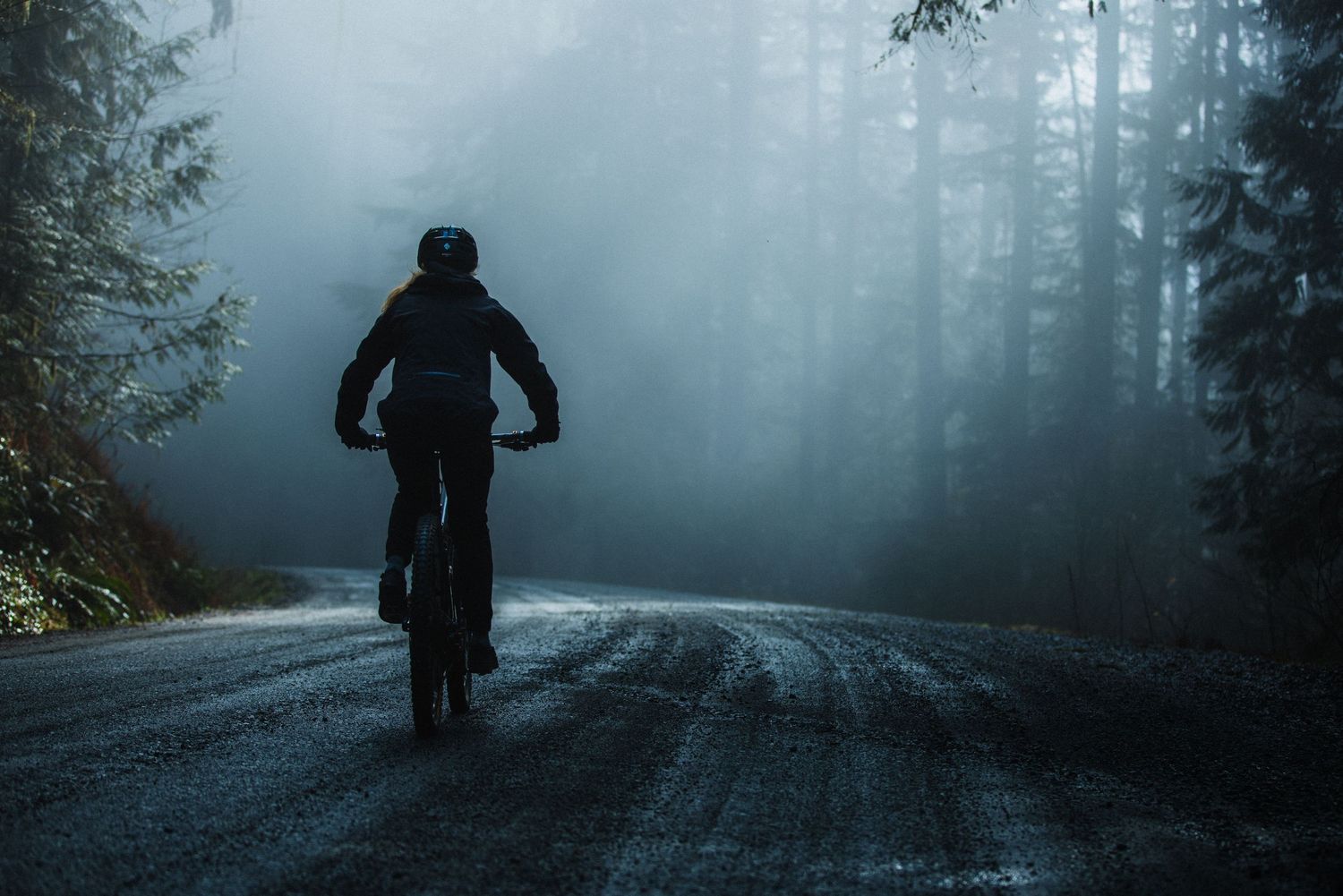 Mountain biker riding a muddy trail through a lush green forest on a full-suspension bike.