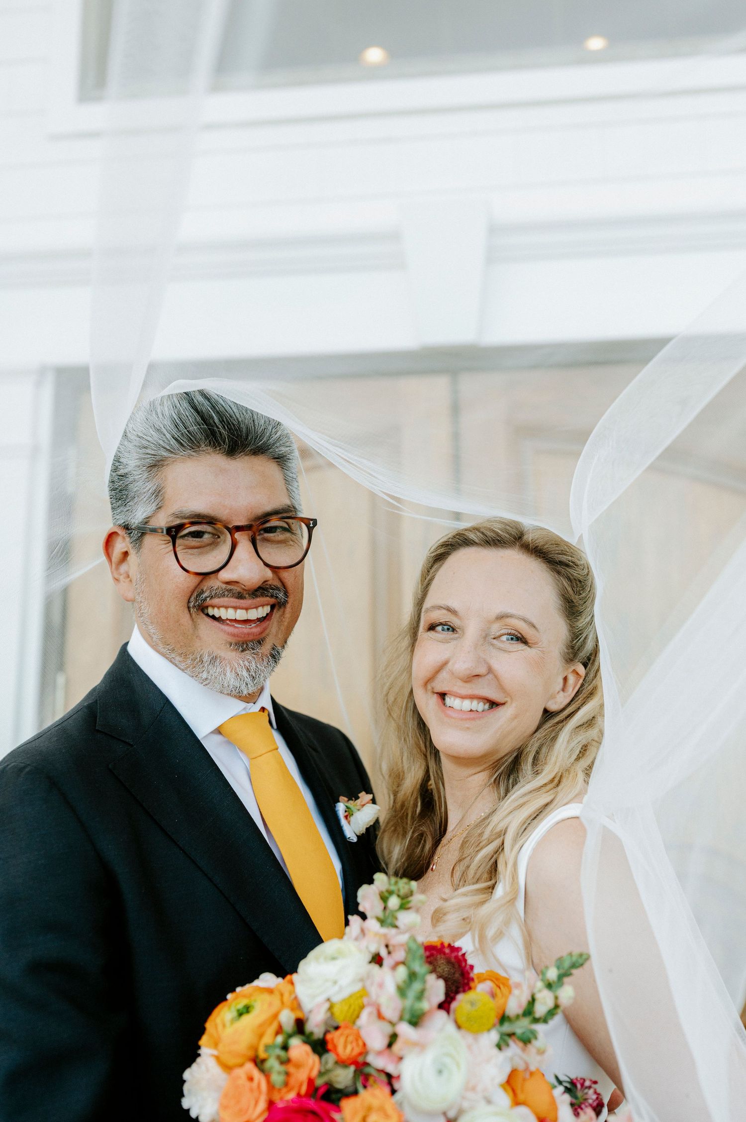 Smiling bride and groom posing together on their wedding day, groom wearing yellow tie and glasses, bride holding colorful bouquet.