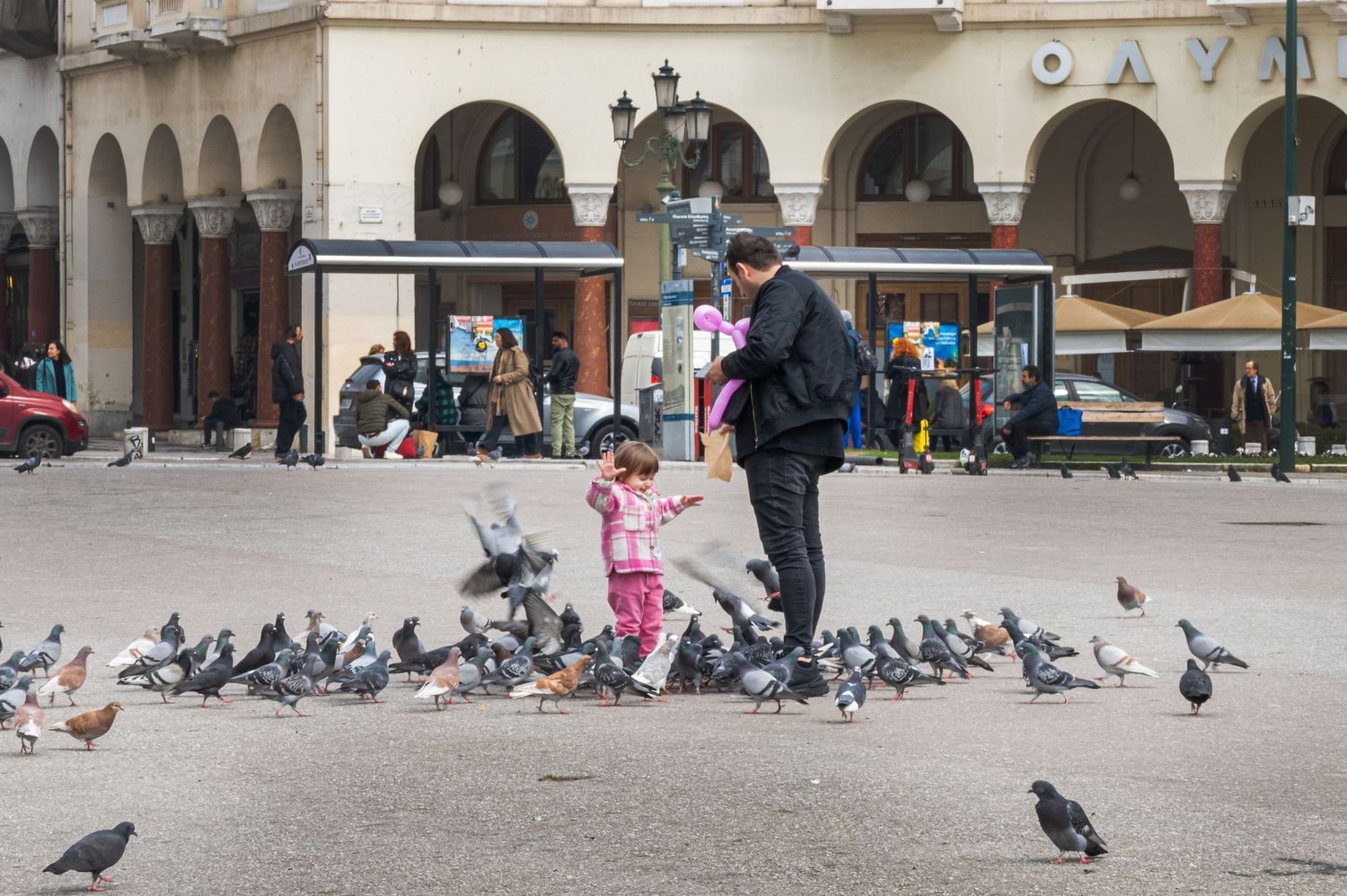 Adult and child feeding pigeons in a busy European town square with historic arcade buildings in the background.