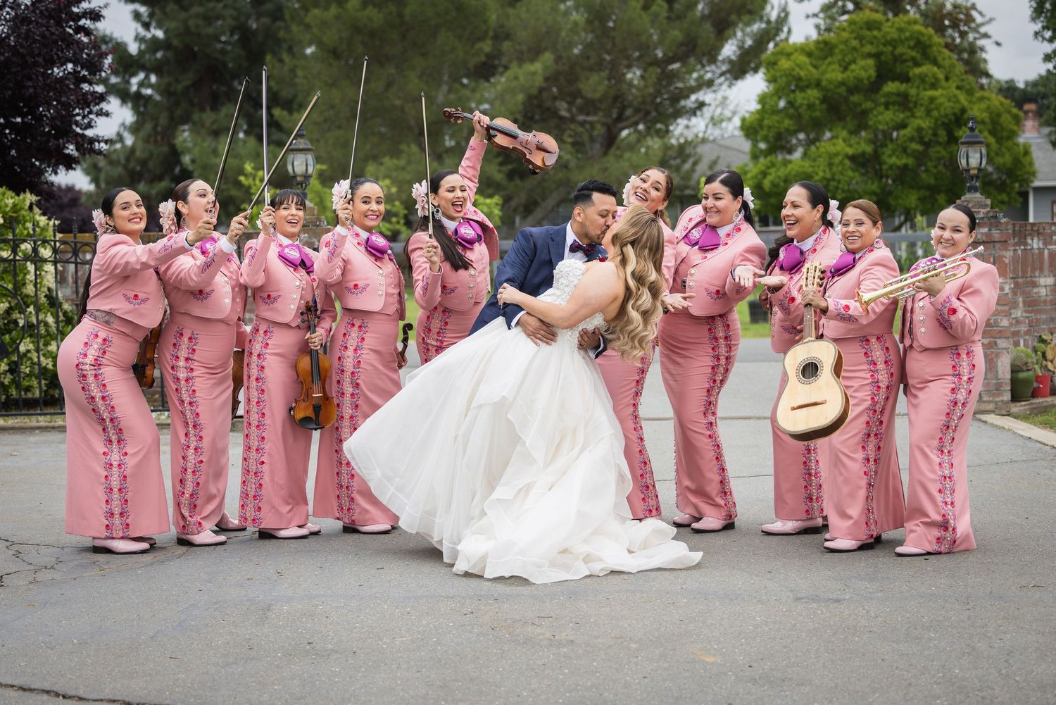Bride and groom share a romantic dip kiss surrounded by an all-female mariachi band in pink traditional outfits at outdoor wedding.