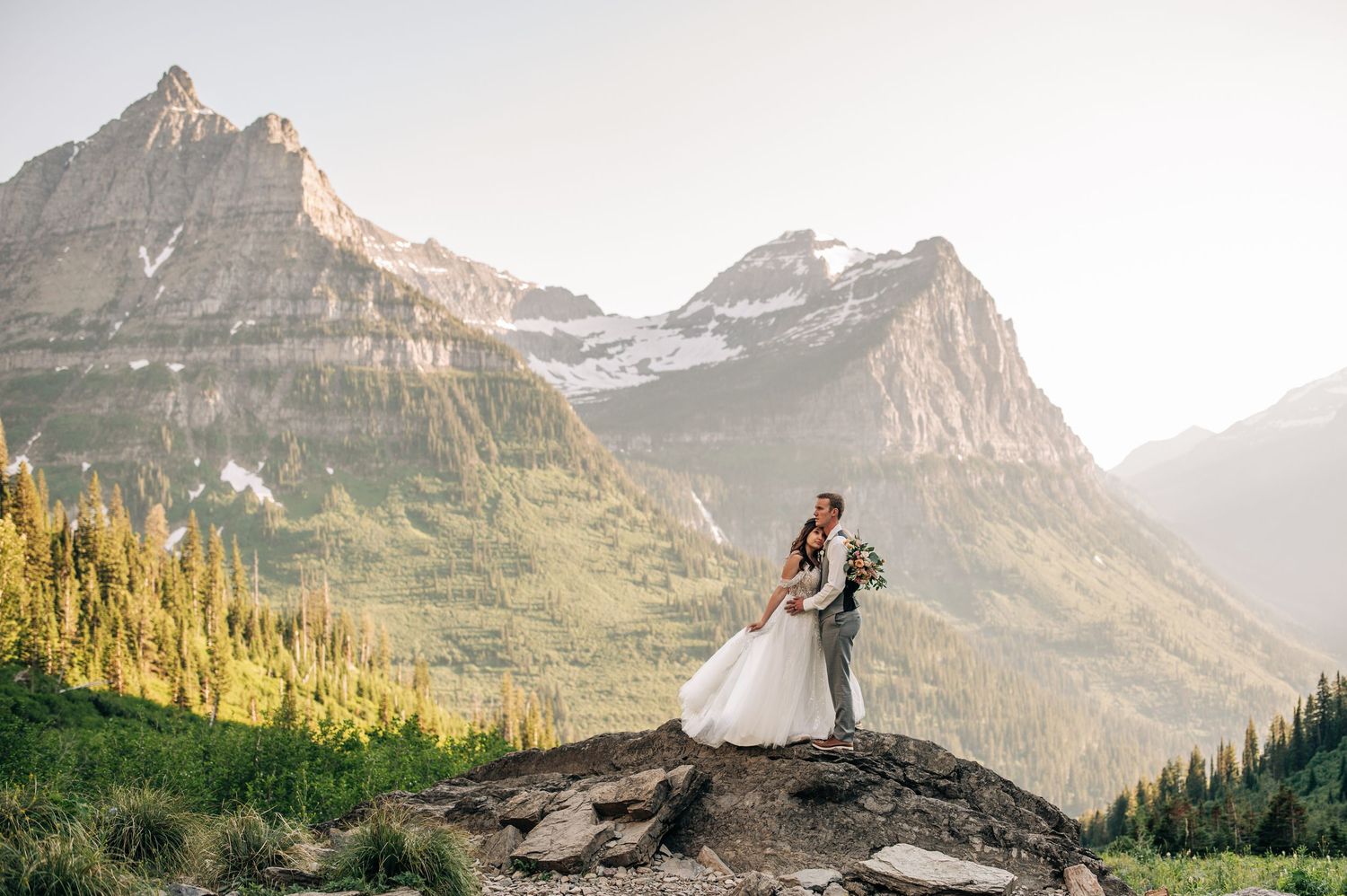 Bride and groom embrace on rocky mountaintop with stunning Glacier National Park peaks and green valleys behind them.