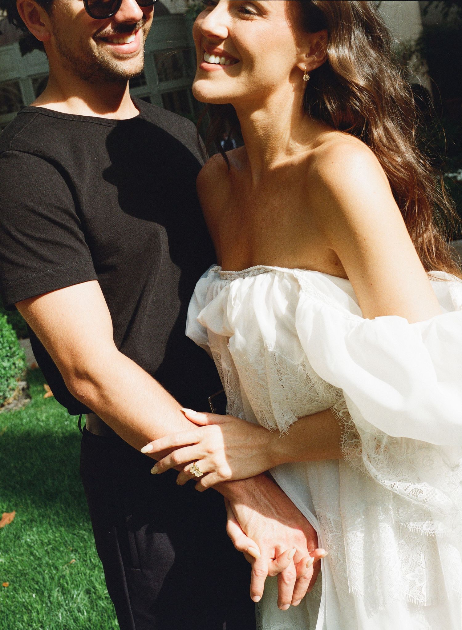 A couple embraces outdoors, woman in white off-shoulder dress and man in black outfit, smiling joyfully.