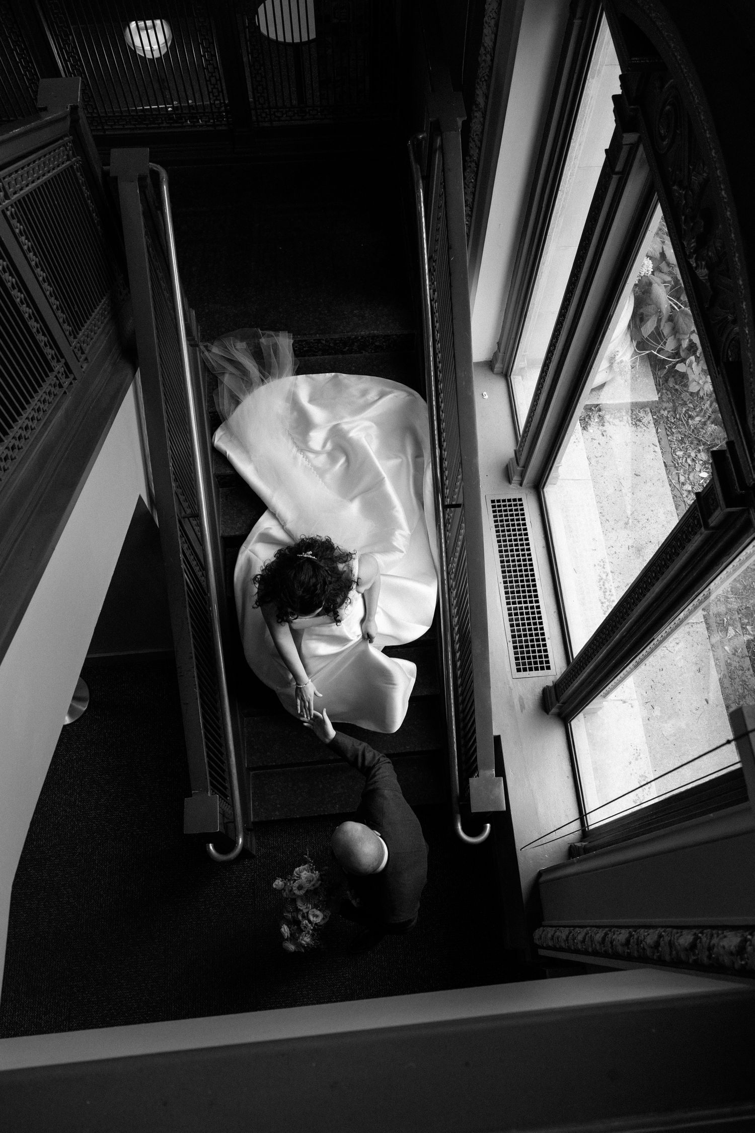 Black and white aerial view of a bride in a flowing white gown descending an elegant staircase with ornate railings.