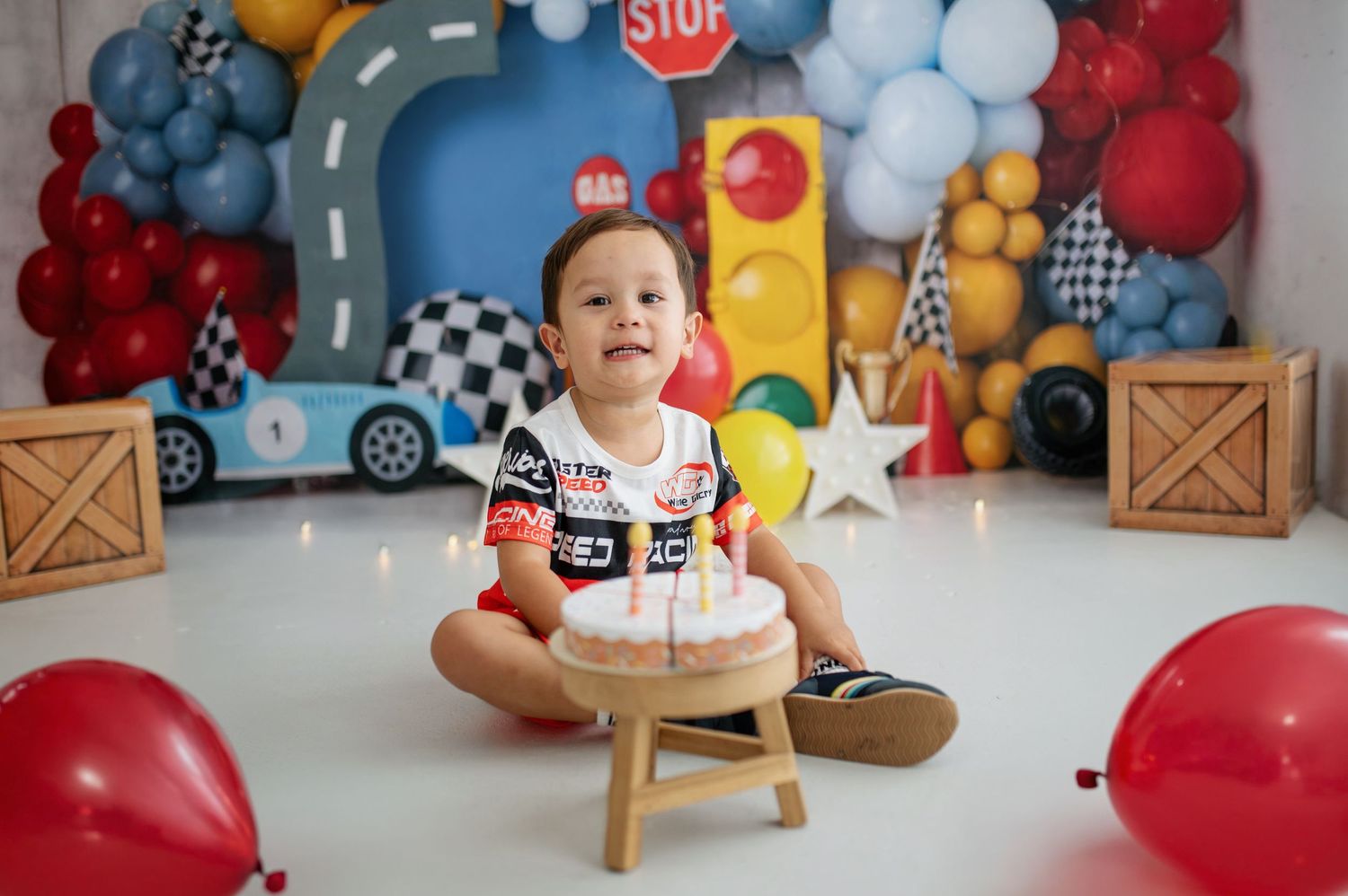 Baby boy in racing outfit sits before a birthday cake, surrounded by colorful balloons and a race car themed backdrop.