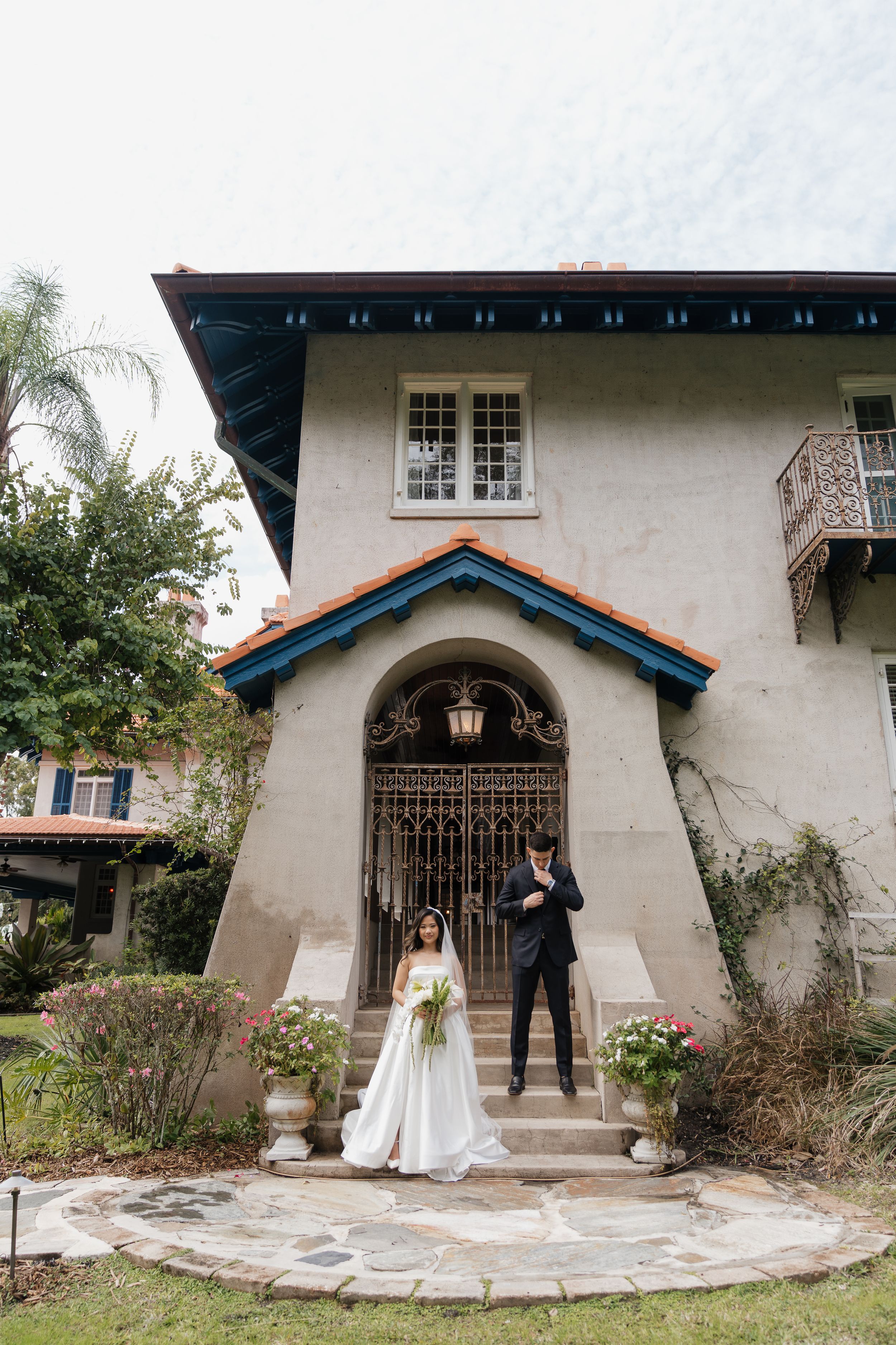 Bride and groom pose on stone steps of elegant Spanish-style villa with ornate iron gate and terracotta roof.