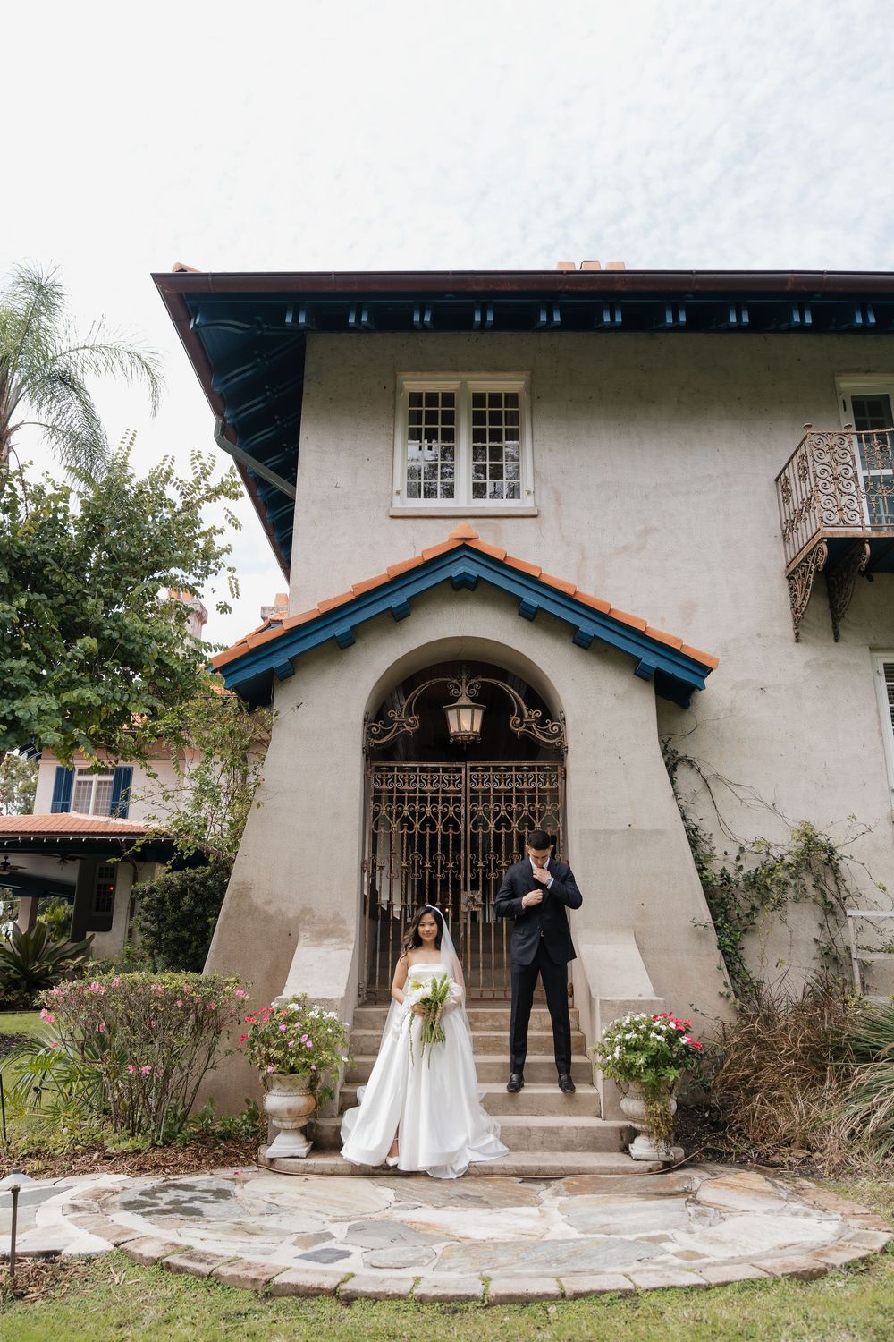Bride and groom pose on stone steps of elegant Spanish-style villa with ornate iron gate and terracotta roof.