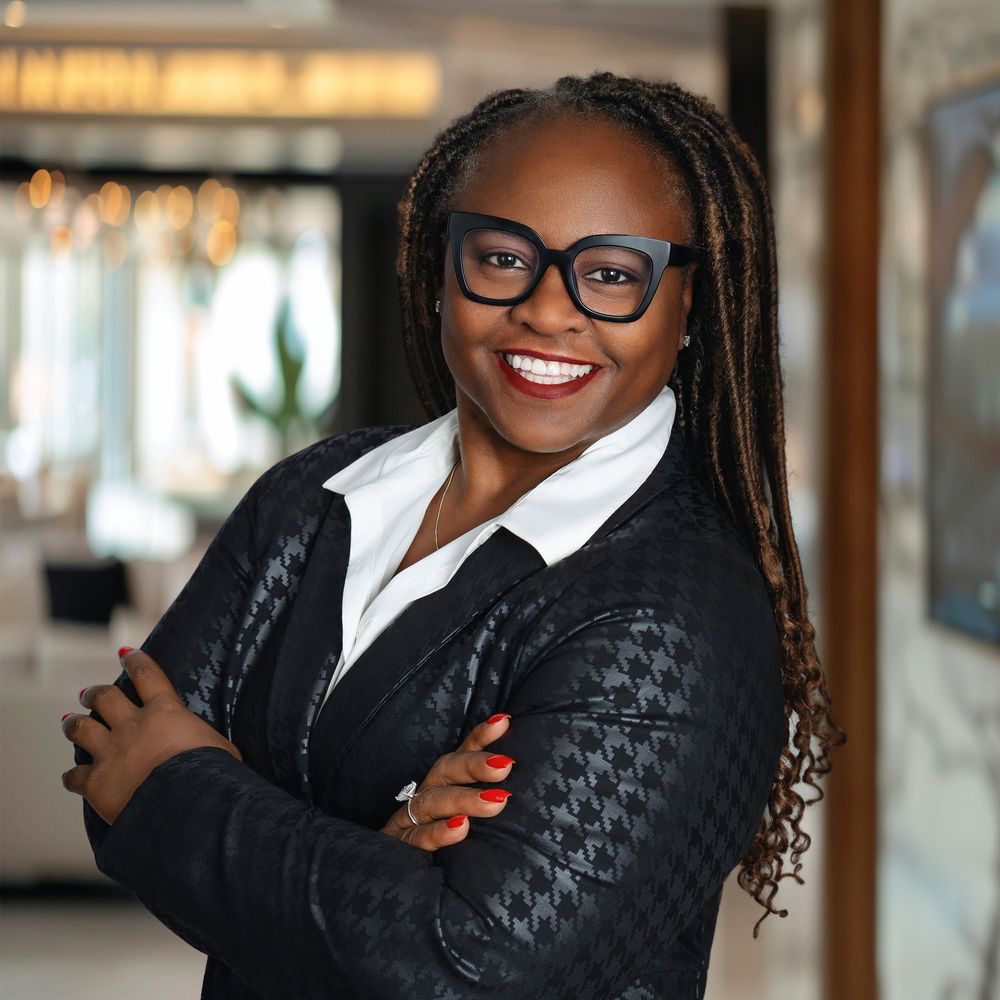 Professional wearing a black blazer and white shirt smiling warmly in a brightly lit office setting.