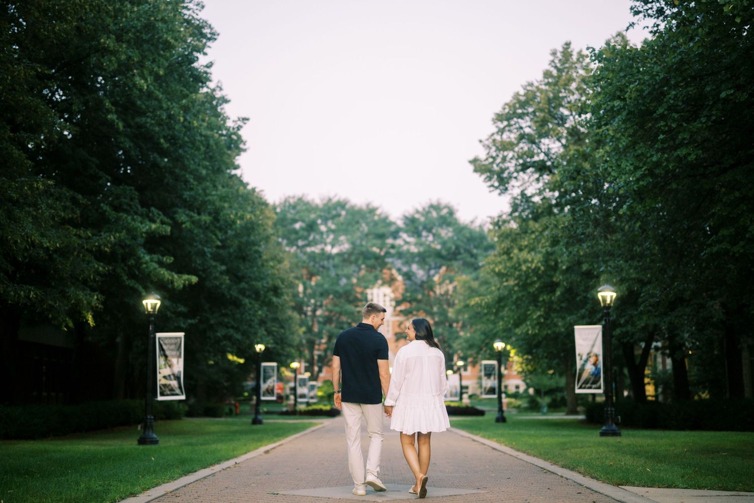 A couple walks hand in hand down a tree-lined path with vintage lampposts at sunset on a college campus.