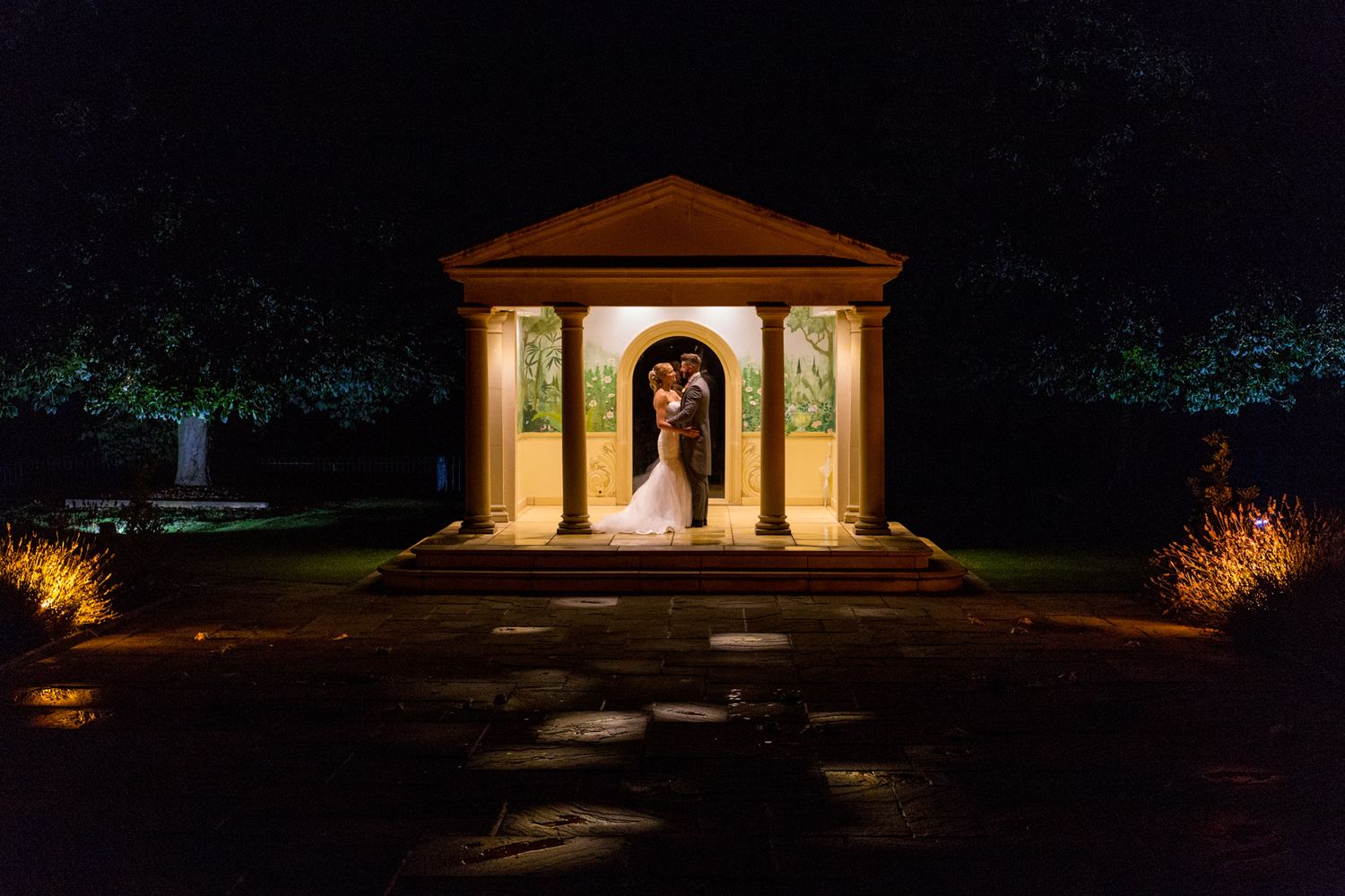 Illuminated gazebo with ornate columns creates romantic nighttime scene in garden with elegant silhouettes standing inside.