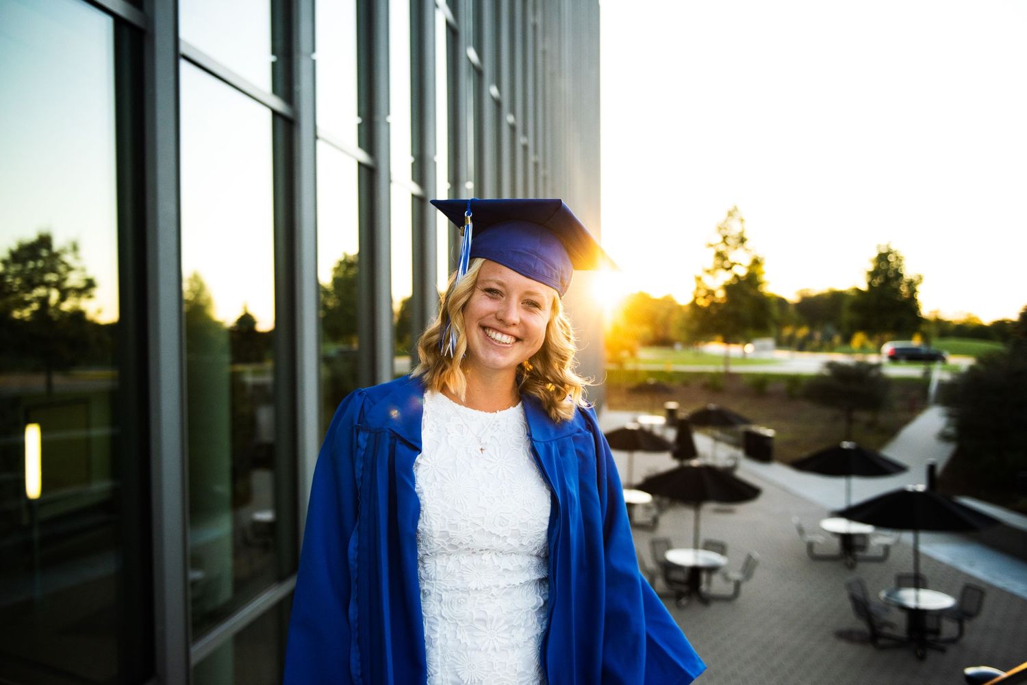 A graduate in blue cap and gown stands outside a modern building at sunset with outdoor seating visible in background.