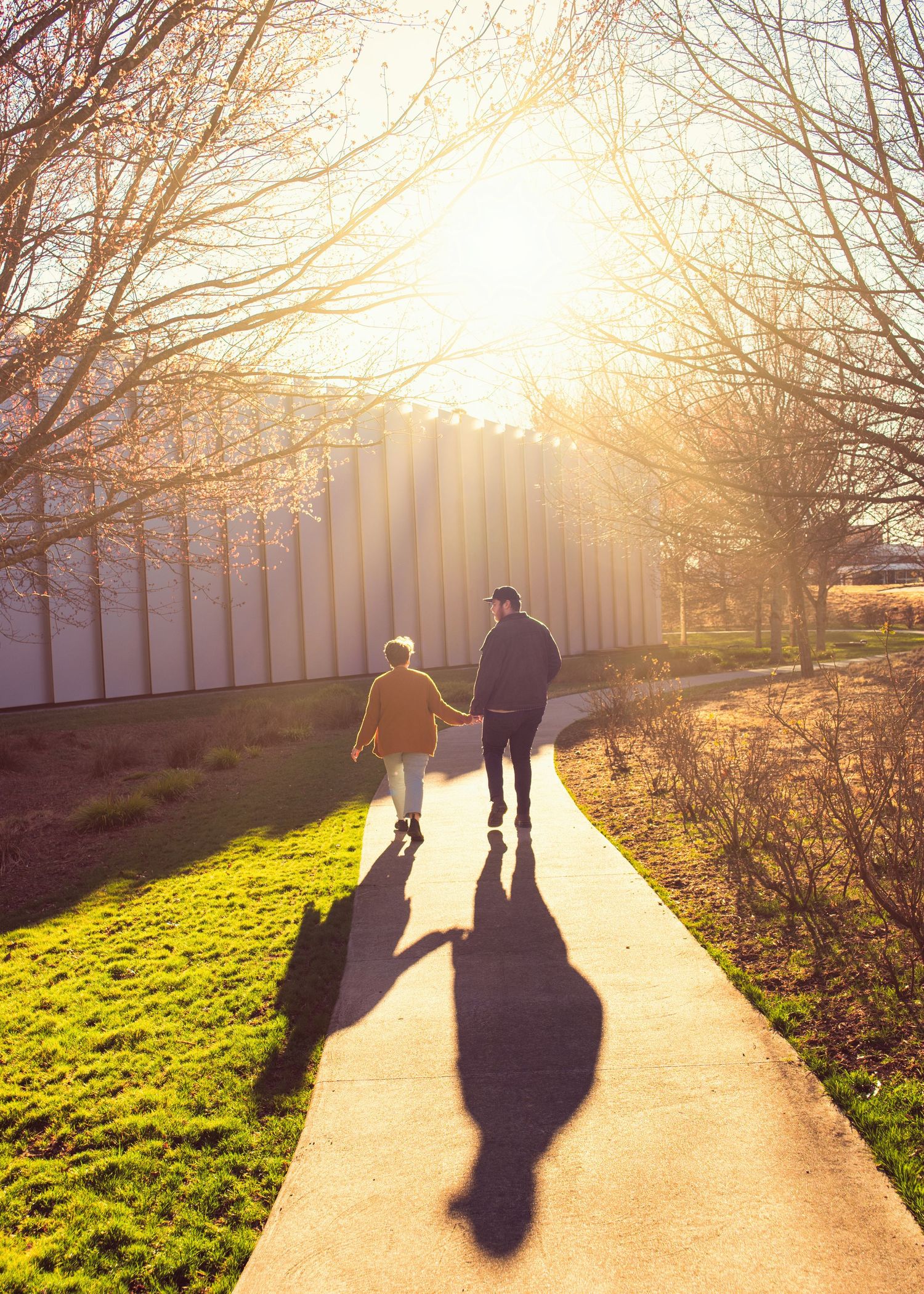 Two people walk along a path at sunset casting long shadows while surrounded by bare trees and grass.