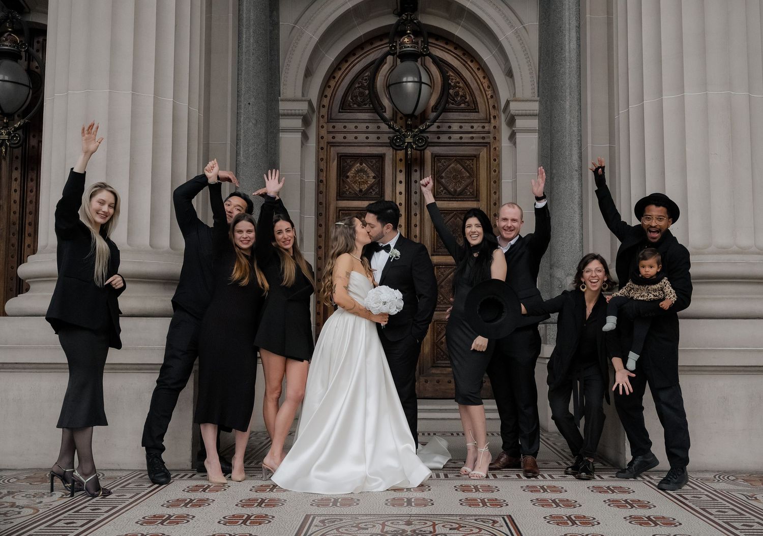 Wedding party celebrates with arms raised outside historic building with grand columns.