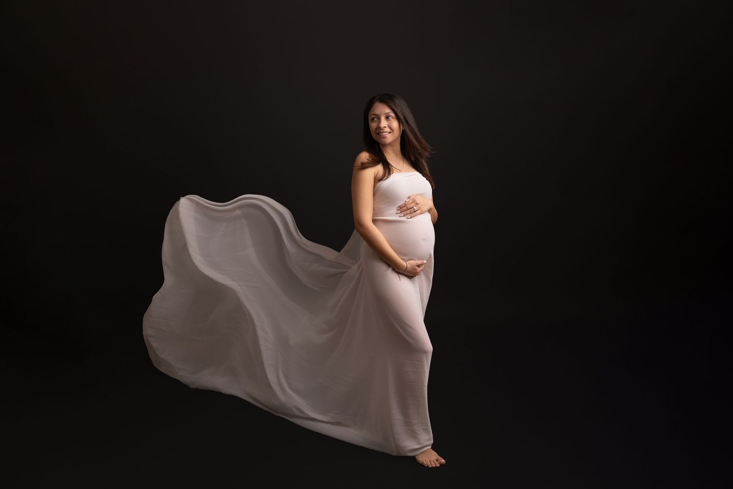 Elegant maternity photo with flowing white dress billowing against dark background in studio setting.