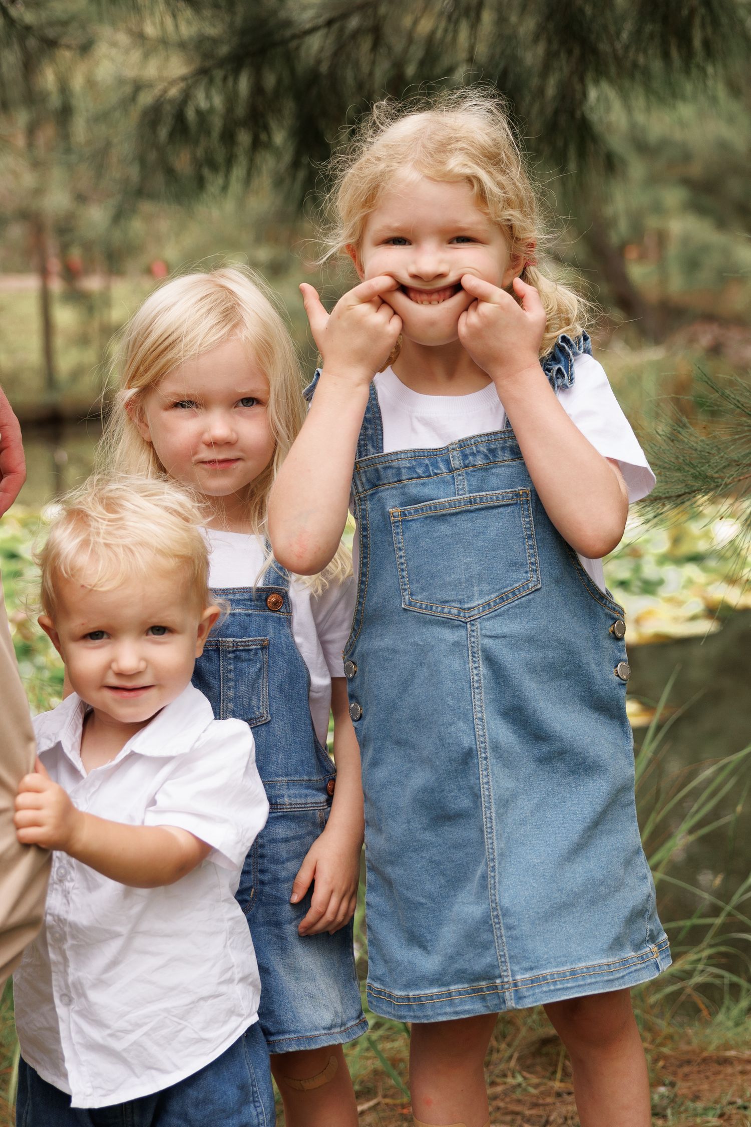 Three young children wearing casual denim and white outfits pose together outdoors among greenery.