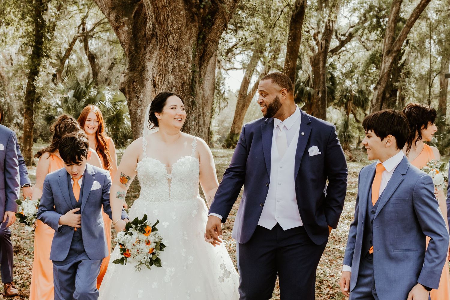 Wedding party walking and posing together outdoors beneath beautiful tree canopy in navy blue and white attire. orange bridesmaid dresses