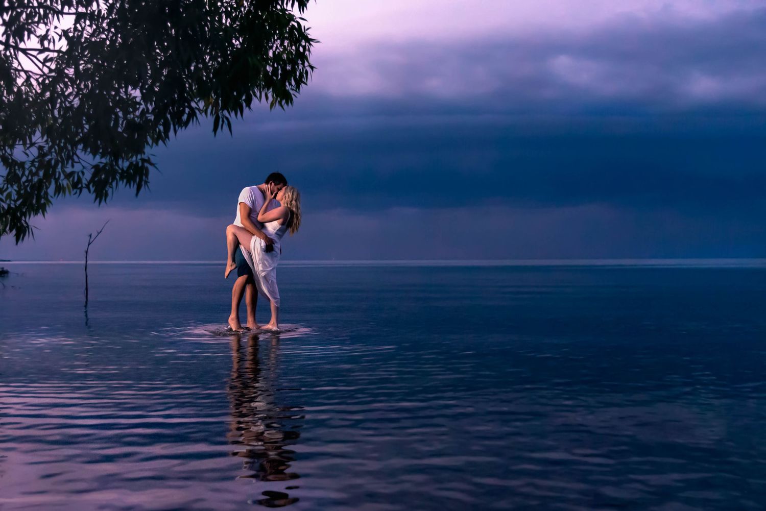 Two people share an intimate moment while standing in shallow reflective water against a moody purple sunset sky.
