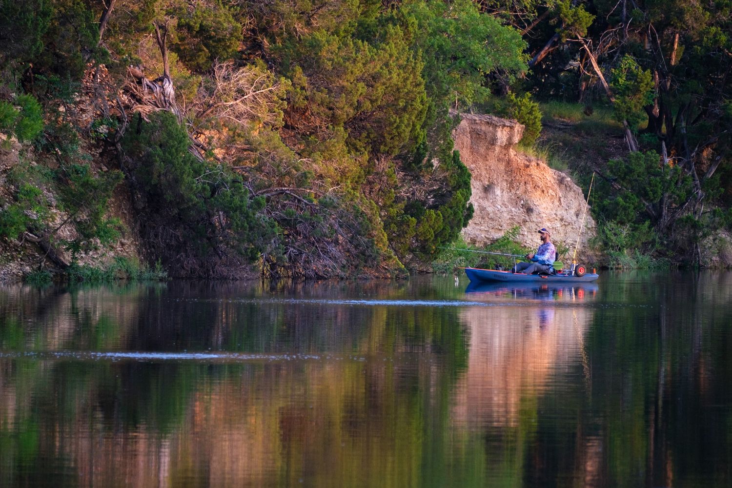 Person fishing from a small boat on a calm reflective river surrounded by lush green cliffs.