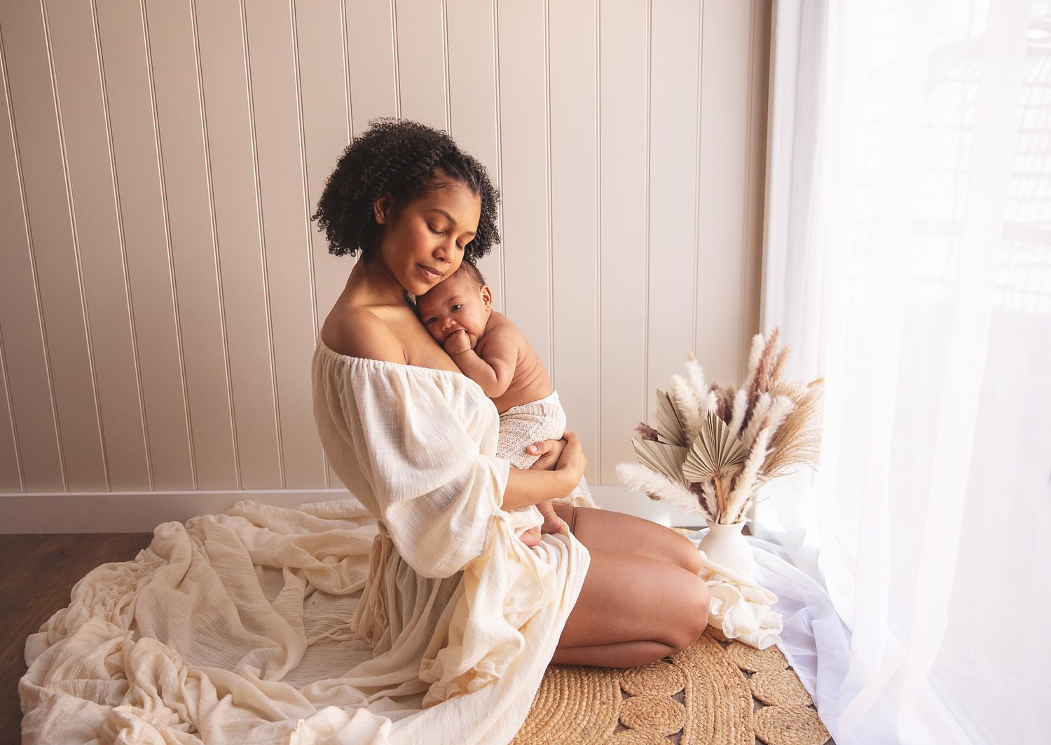 A woman in a flowing white dress sits on the floor near a window with dried pampas grass decor. During a newborn photoshoot in London